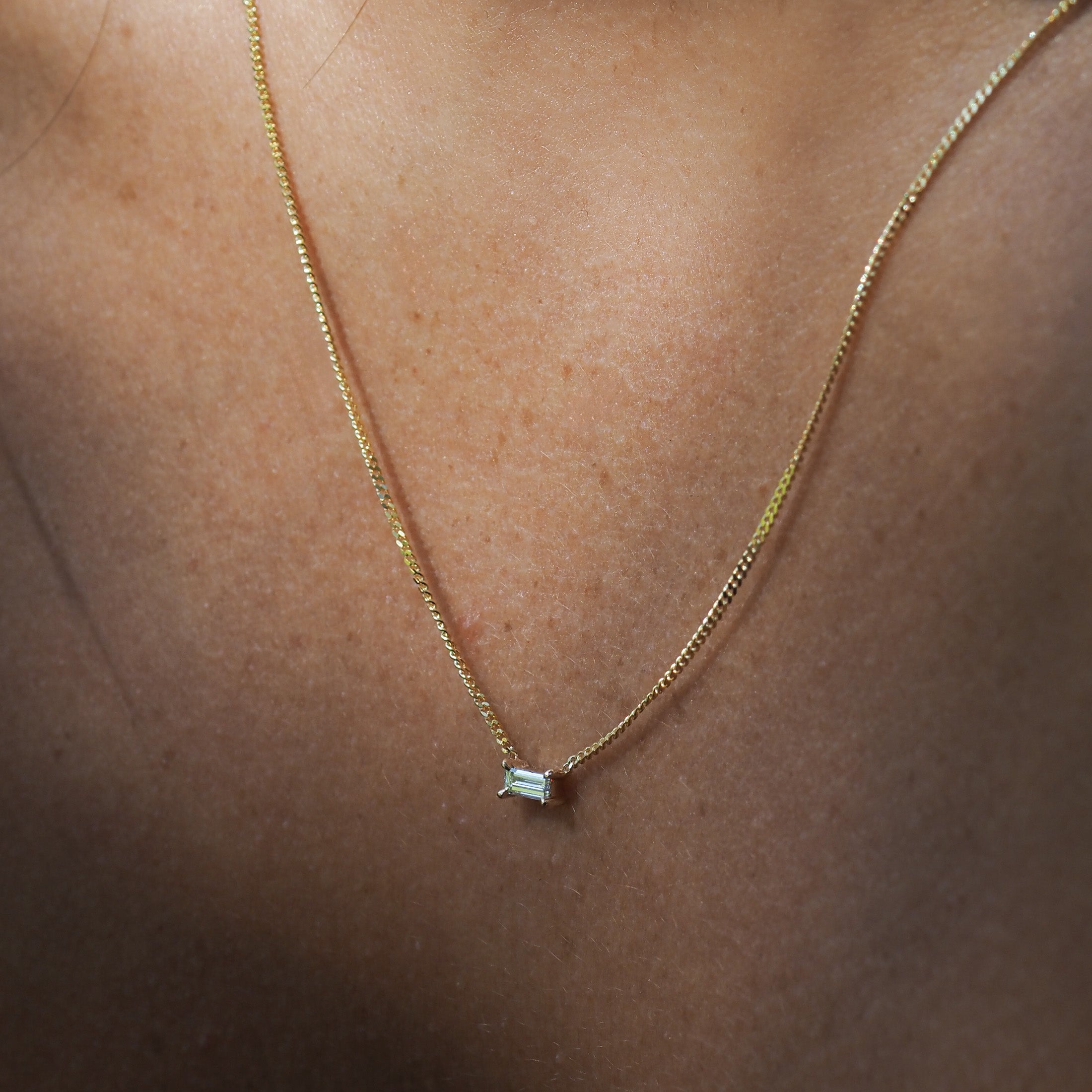 Gold chain with a small emerald-cut diamond pendant on a person's neck, showcasing delicate luxury jewellery in soft lighting.