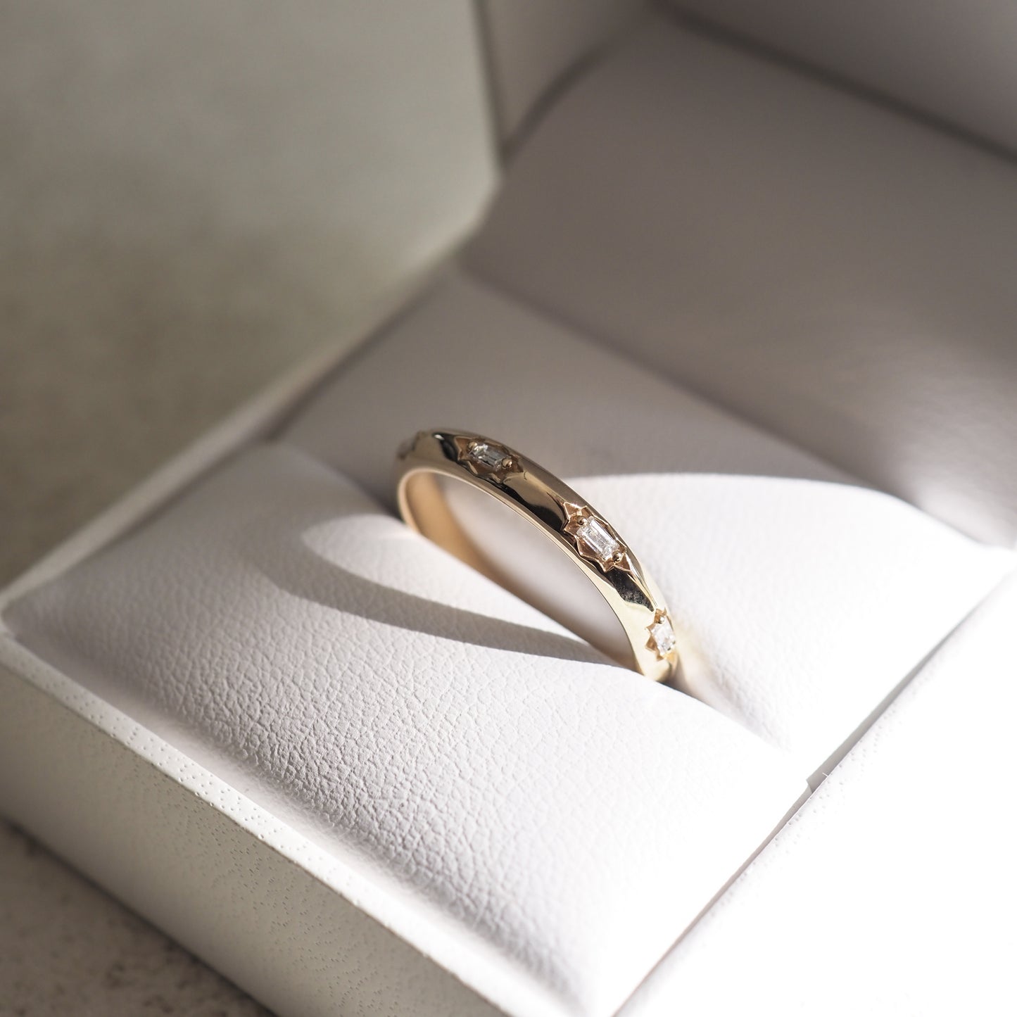Gold band with star-shaped diamond inlays in a white leather jewellery box, illuminated by soft sunlight.