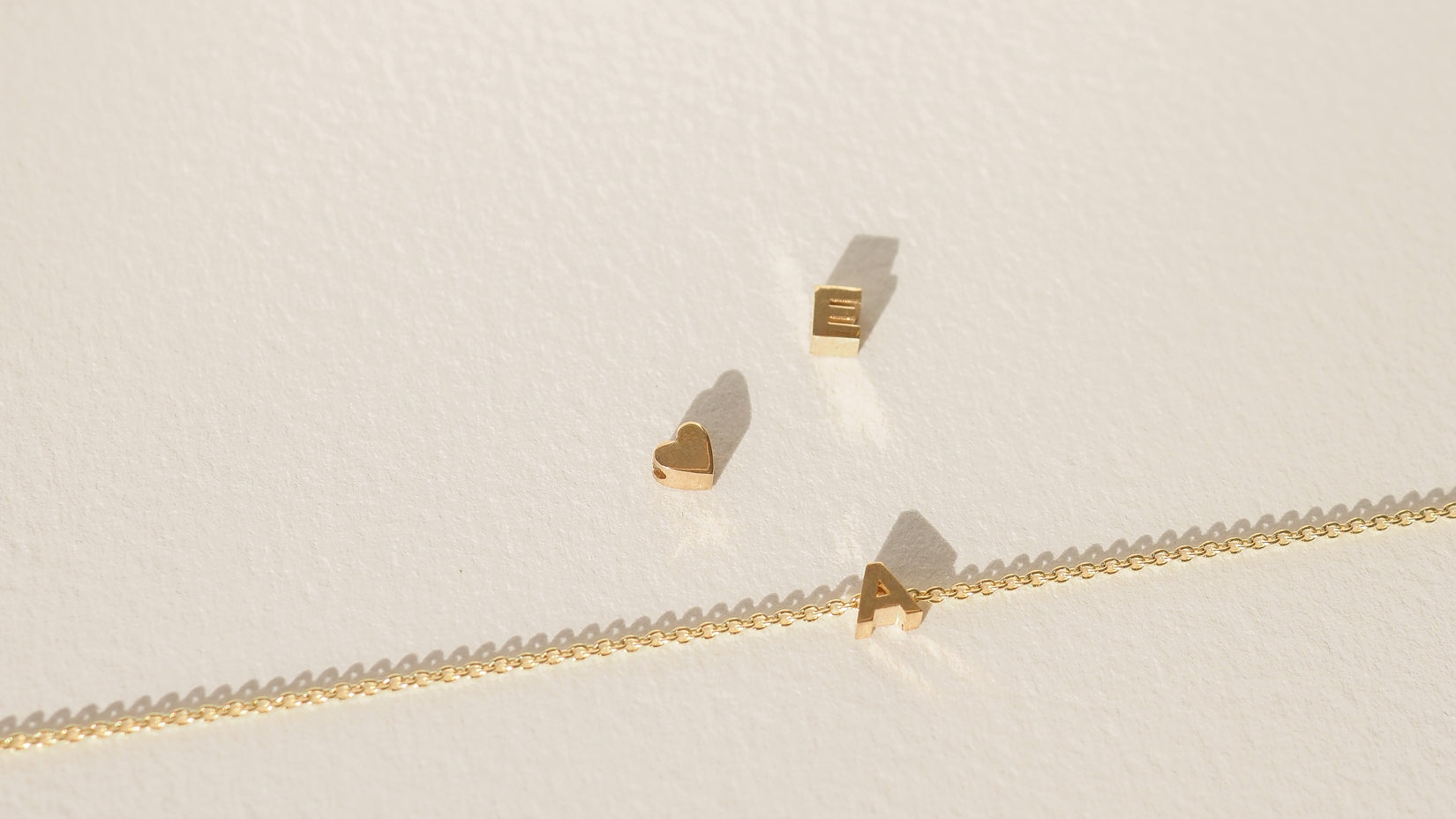 Gold chain with letter and heart charms on a light background; minimalist luxury jewellery aesthetic.