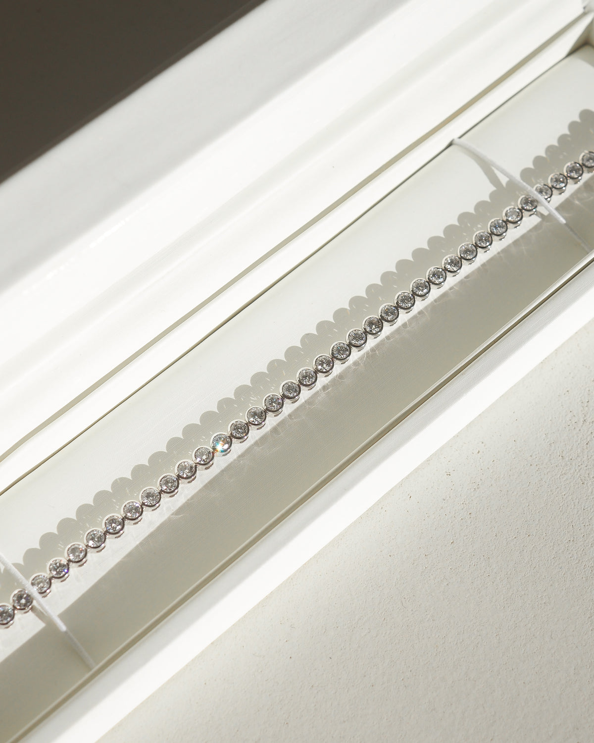 A close-up of a delicate white gold bezel tennis bracelet displayed on a white curved stand.