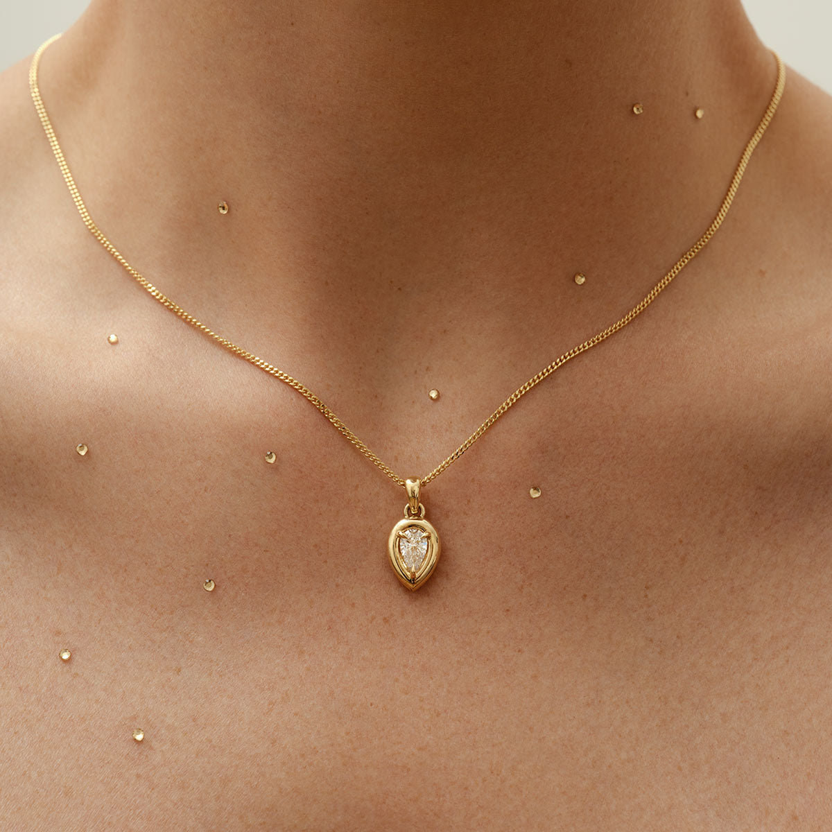 Gold necklace with a teardrop pendant featuring a diamond, against a skin tone backdrop adorned with small gold dots. Luxurious jewellery.