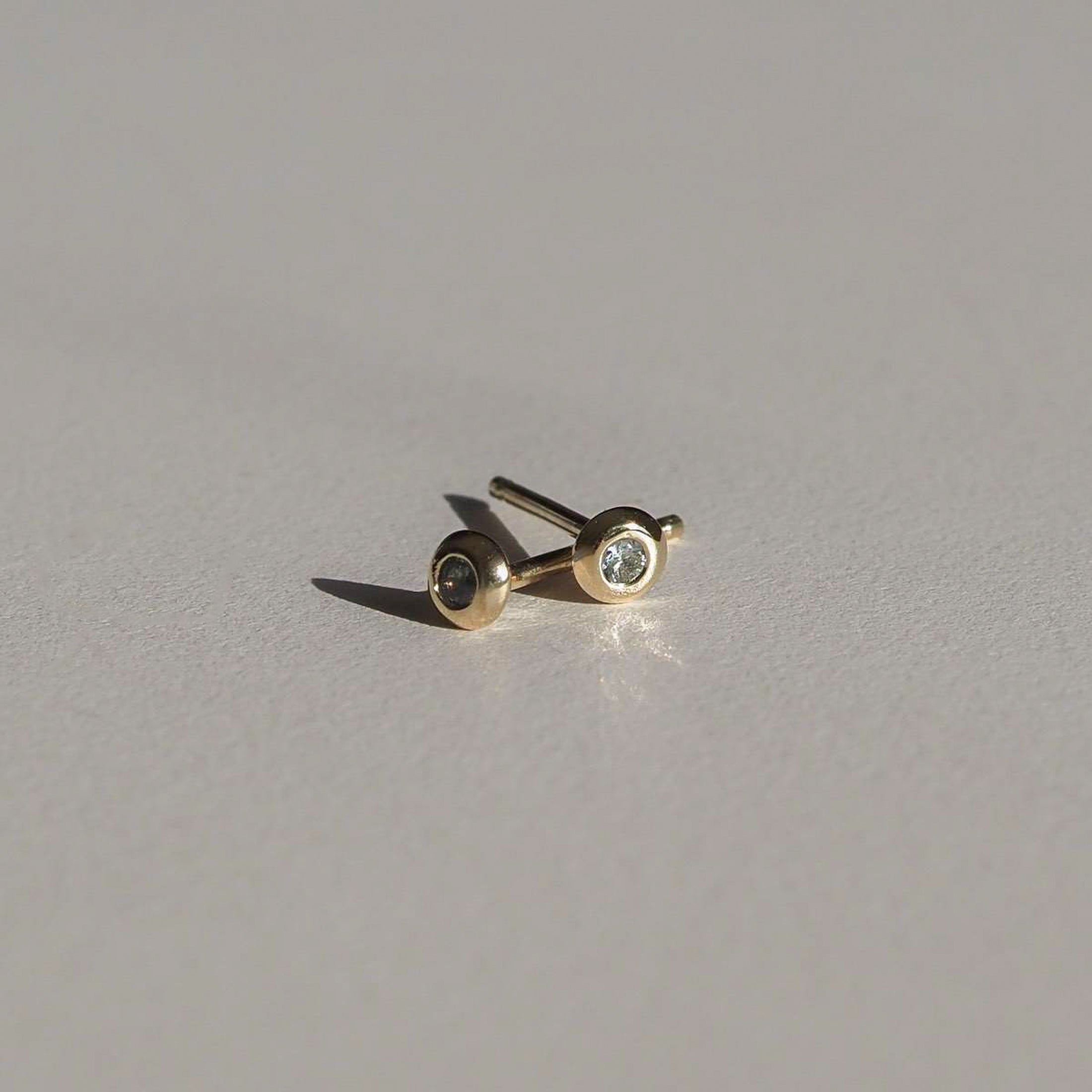 Gold stud earrings with small centred diamonds, elegantly displayed on a light grey surface. Minimalist luxury jewellery.