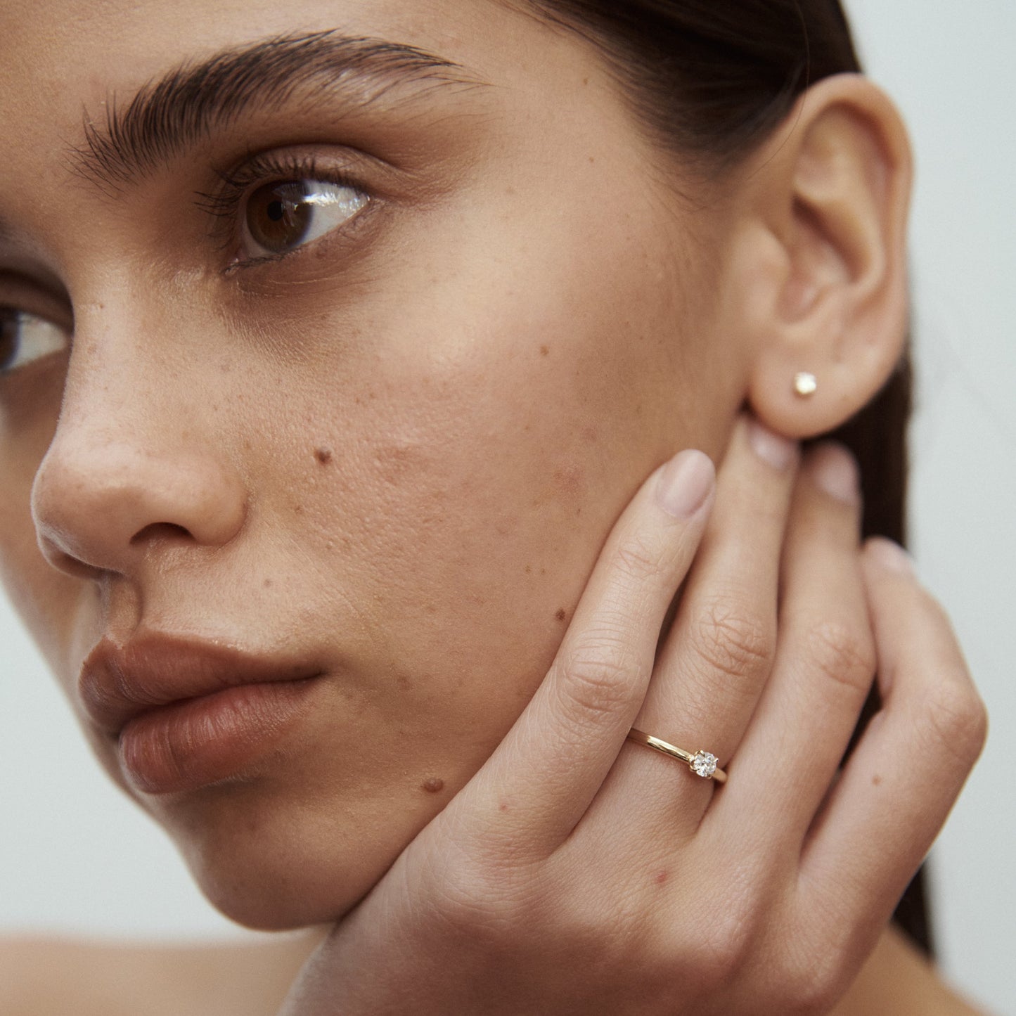 Close-up of a woman's face highlighting a gold ring with a diamond on her finger and a small stud earring. Luxurious and elegant jewellery.
