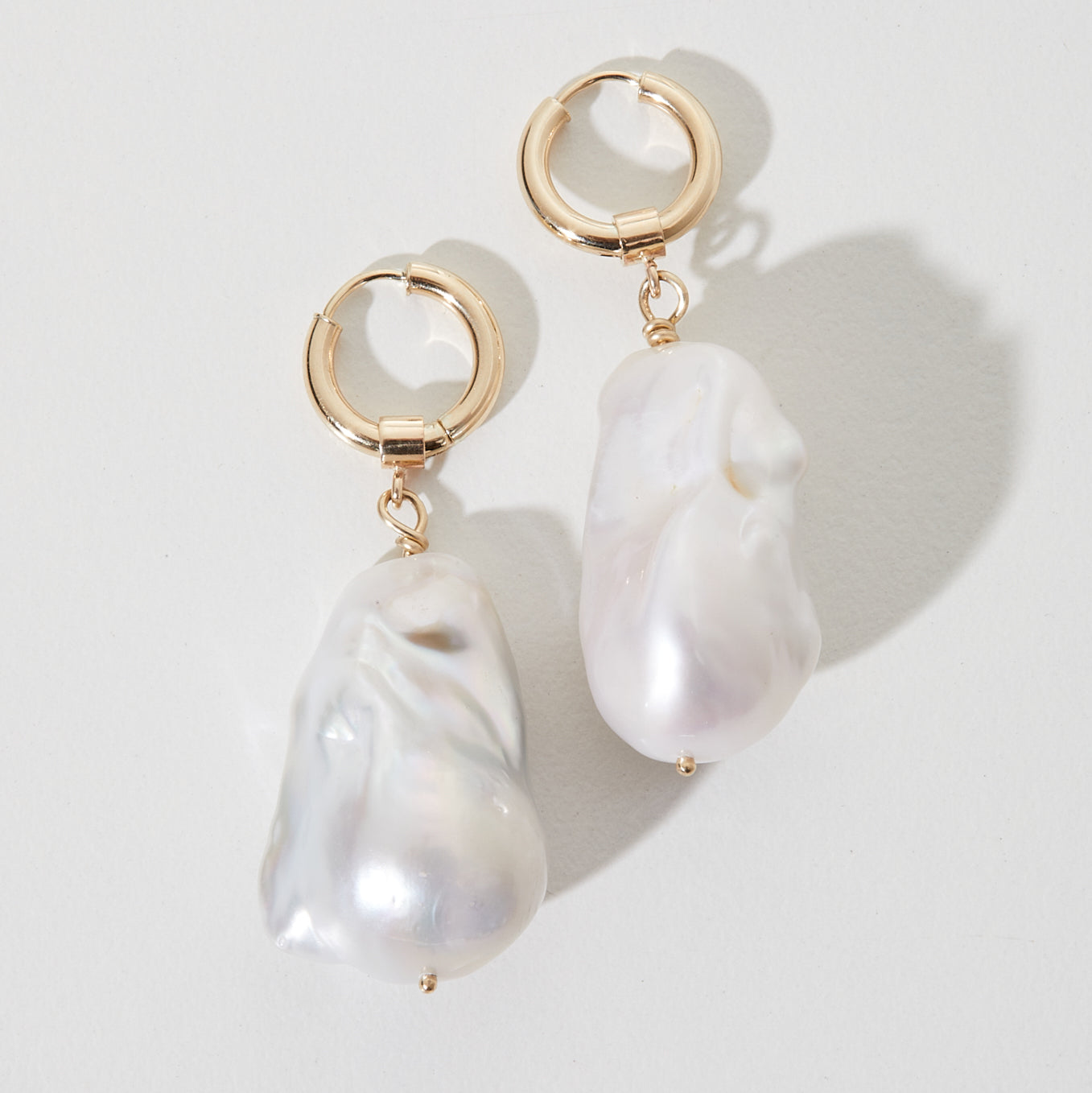 Gold hoop earrings with large, irregular baroque pearls, casting soft shadows on a light grey background. Luxury jewellery design.