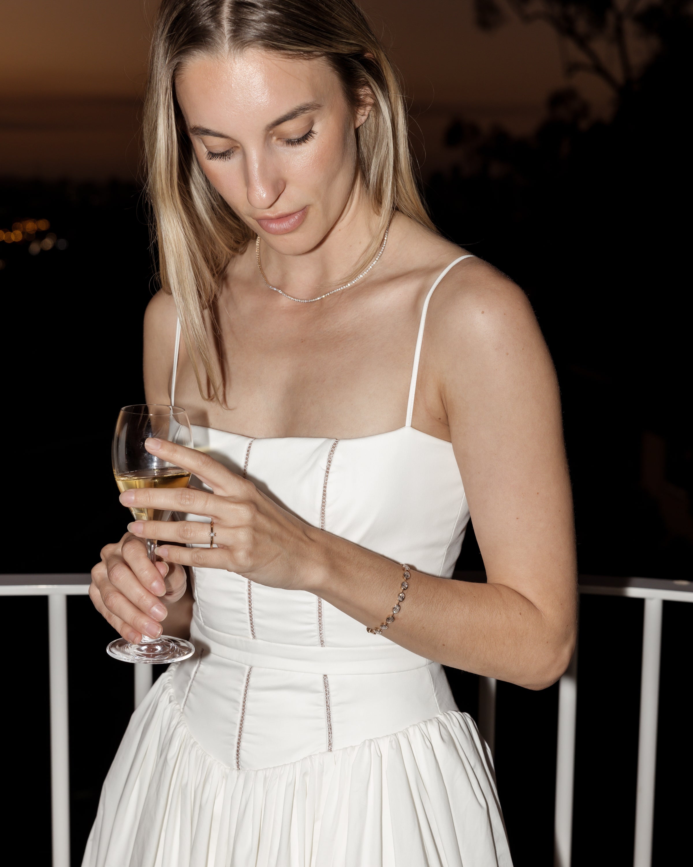 Woman in a white dress holding a wine glass, wearing a delicate necklace and bracelet, standing on a balcony at dusk. Luxury jewellery.
