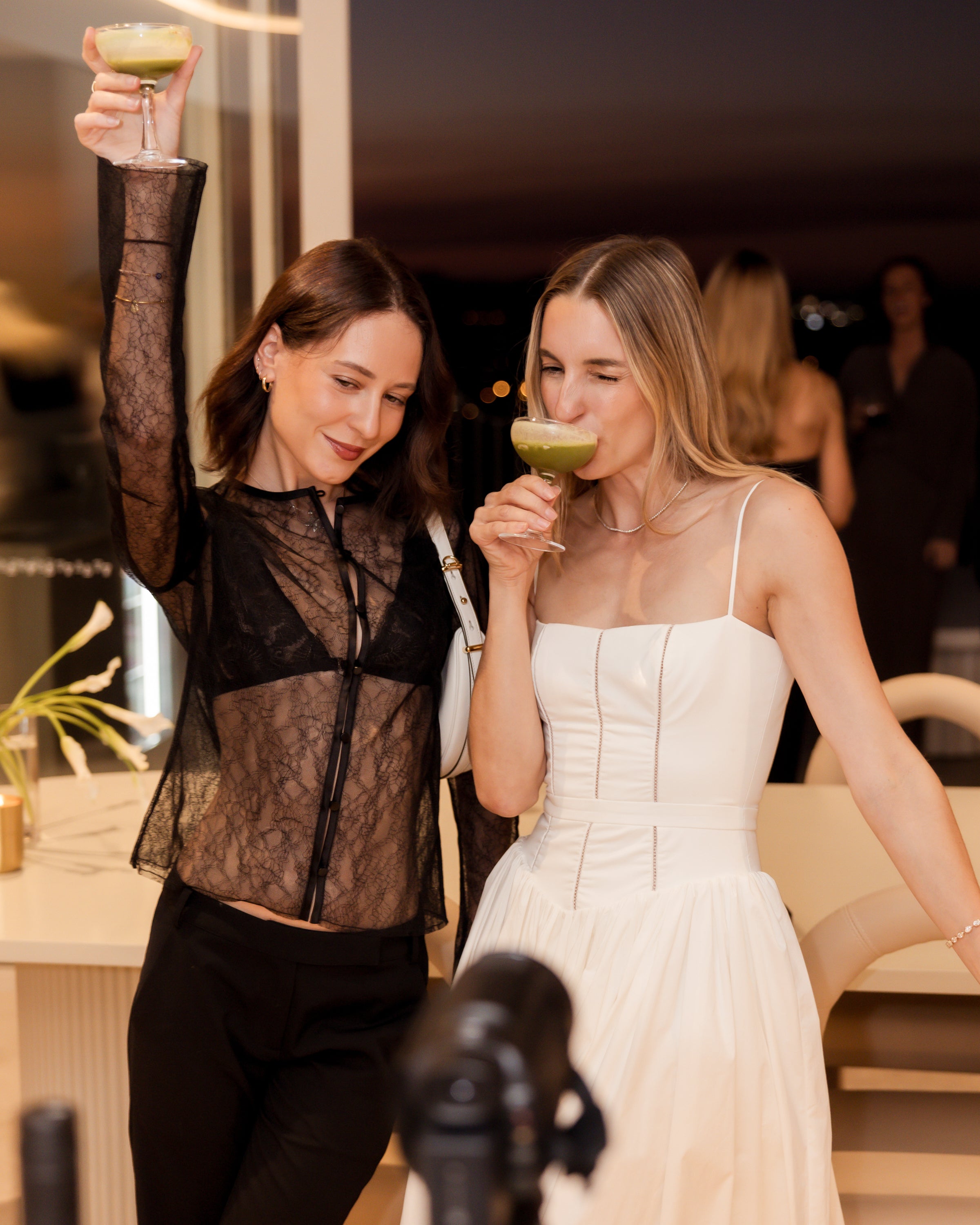 Two women at a luxury event, smiling and holding drinks. One wears a sheer lace top, the other a white dress. Elegant jewellery and decor visible.