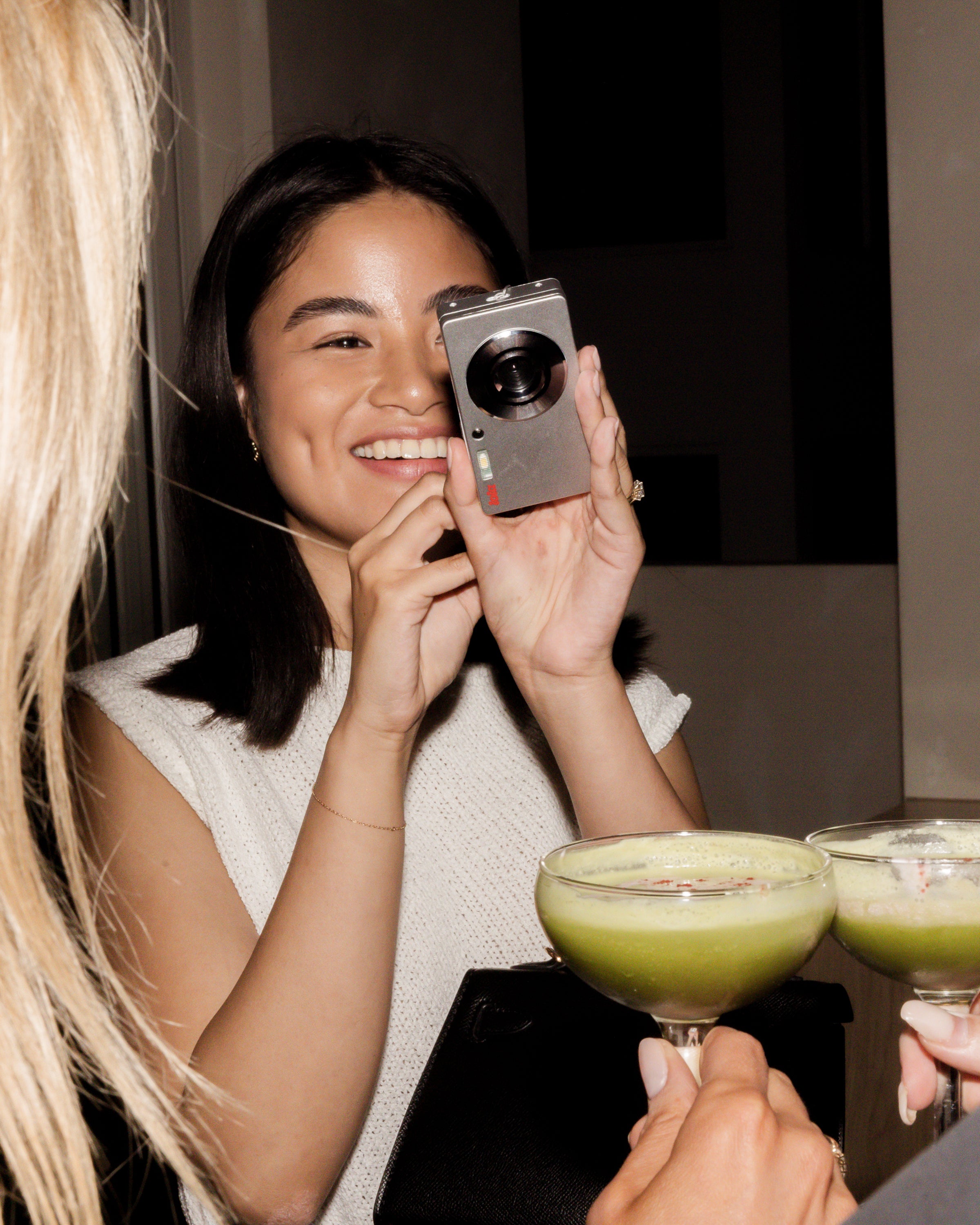Woman in a white top holds a camera, capturing a moment at a party with margarita cocktails. Luxe jewellery subtly accents the scene.