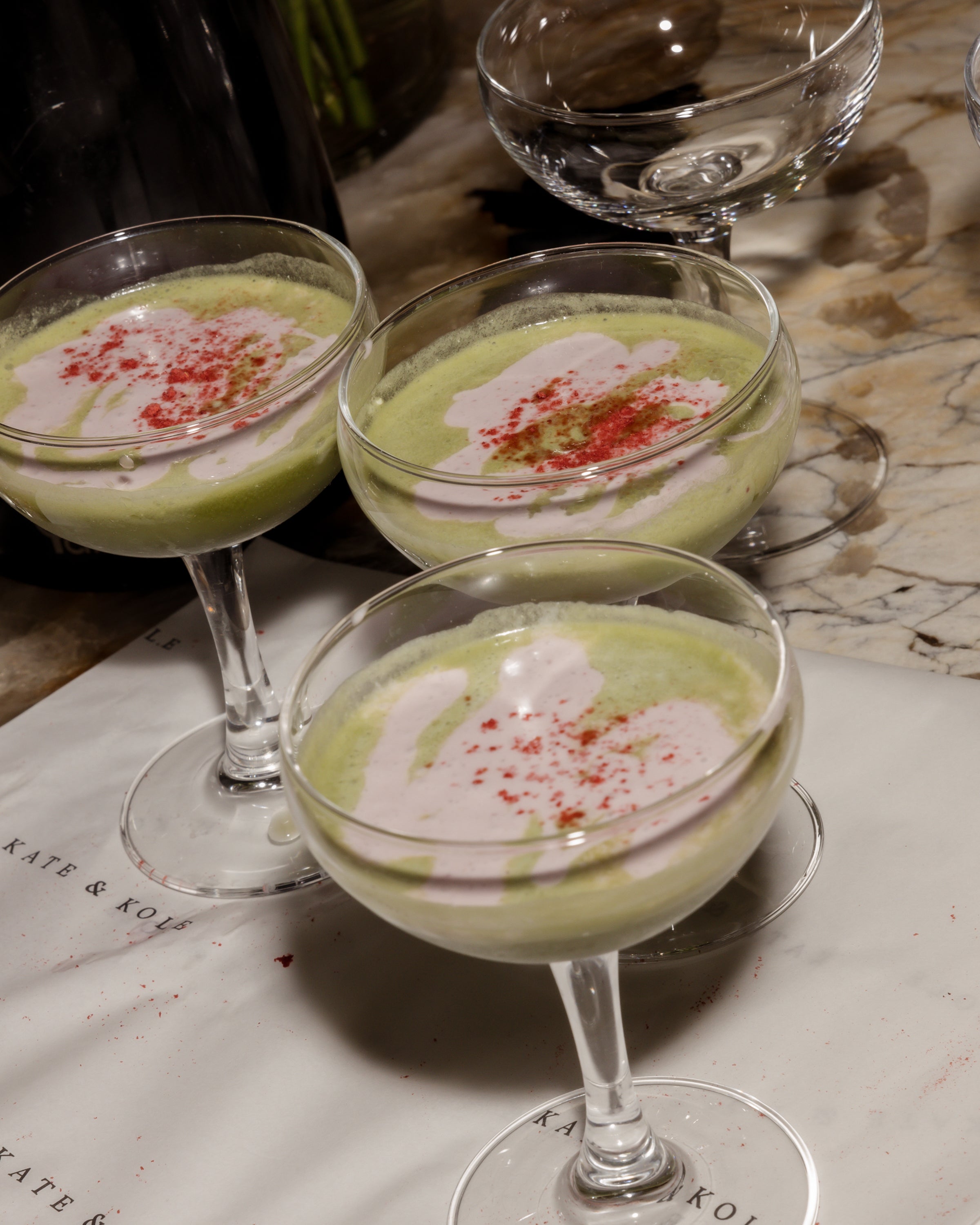 Three elegant cocktail glasses filled with a creamy green beverage, topped with pink drizzle and red sprinkles, on a marbled surface.