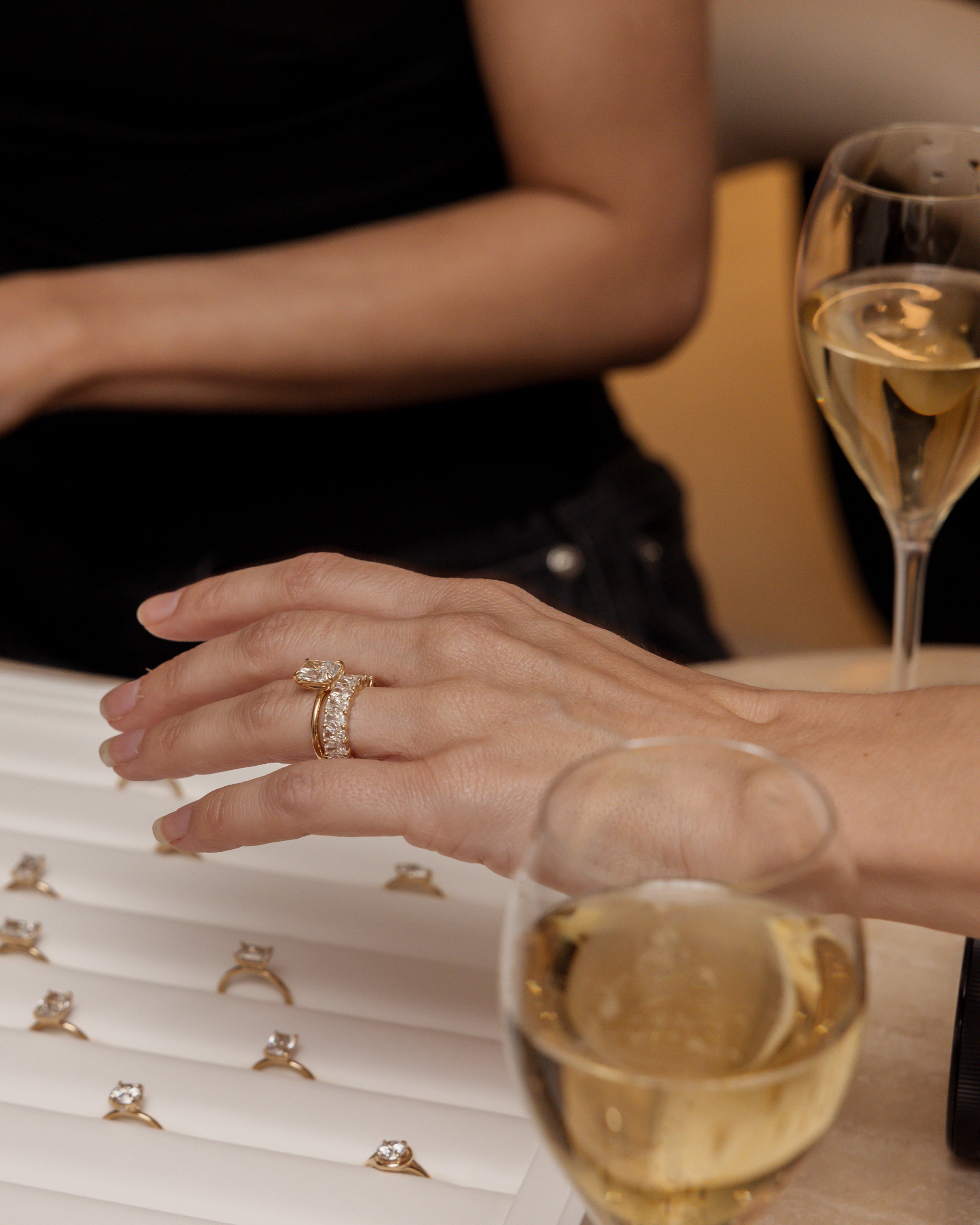A hand trying on a diamond engagement ring from a selection, with champagne glasses in the background, suggesting a luxury jewellery setting.