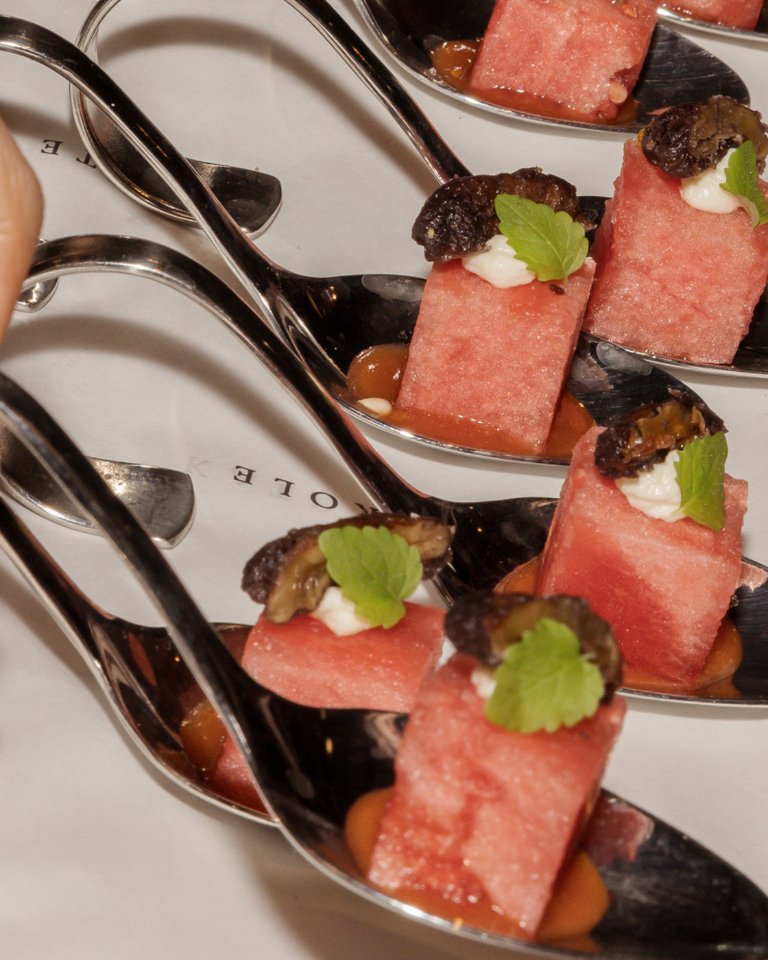 Elegant appetisers on silver spoons, featuring watermelon cubes topped with cream, garnished with a mint leaf and dried fruit, on a white surface.