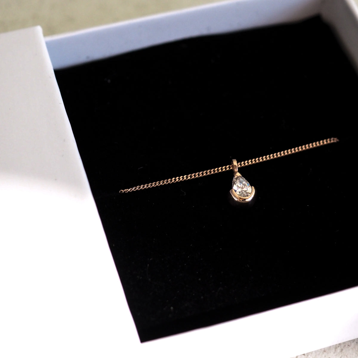 Gold chain with a teardrop diamond pendant in an open white jewellery box, displayed on a black velvet lining.