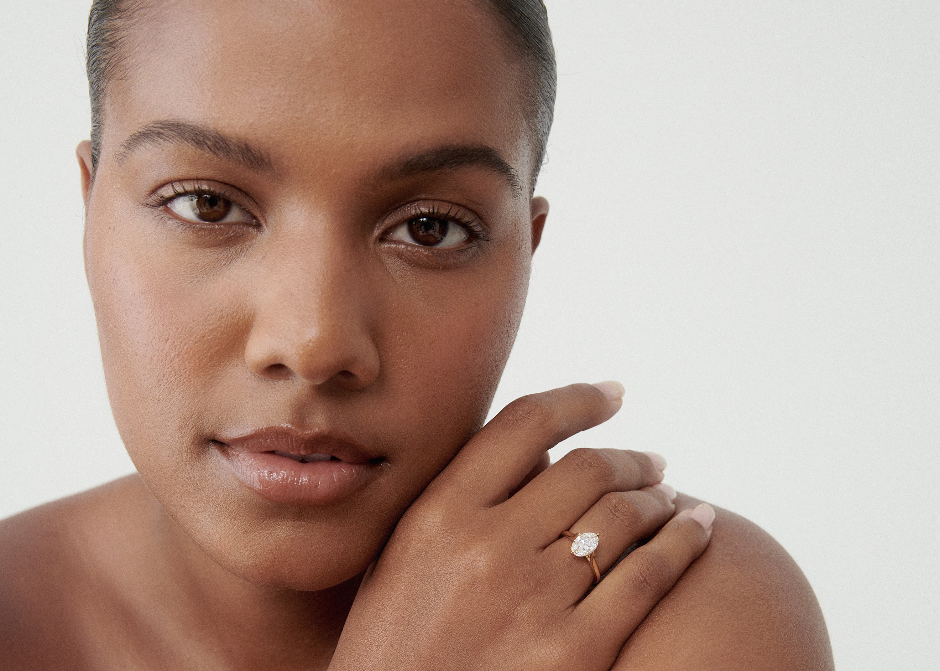 Close-up of a woman with a sleek hairstyle, showcasing a luxurious diamond ring on her finger against a simple background.