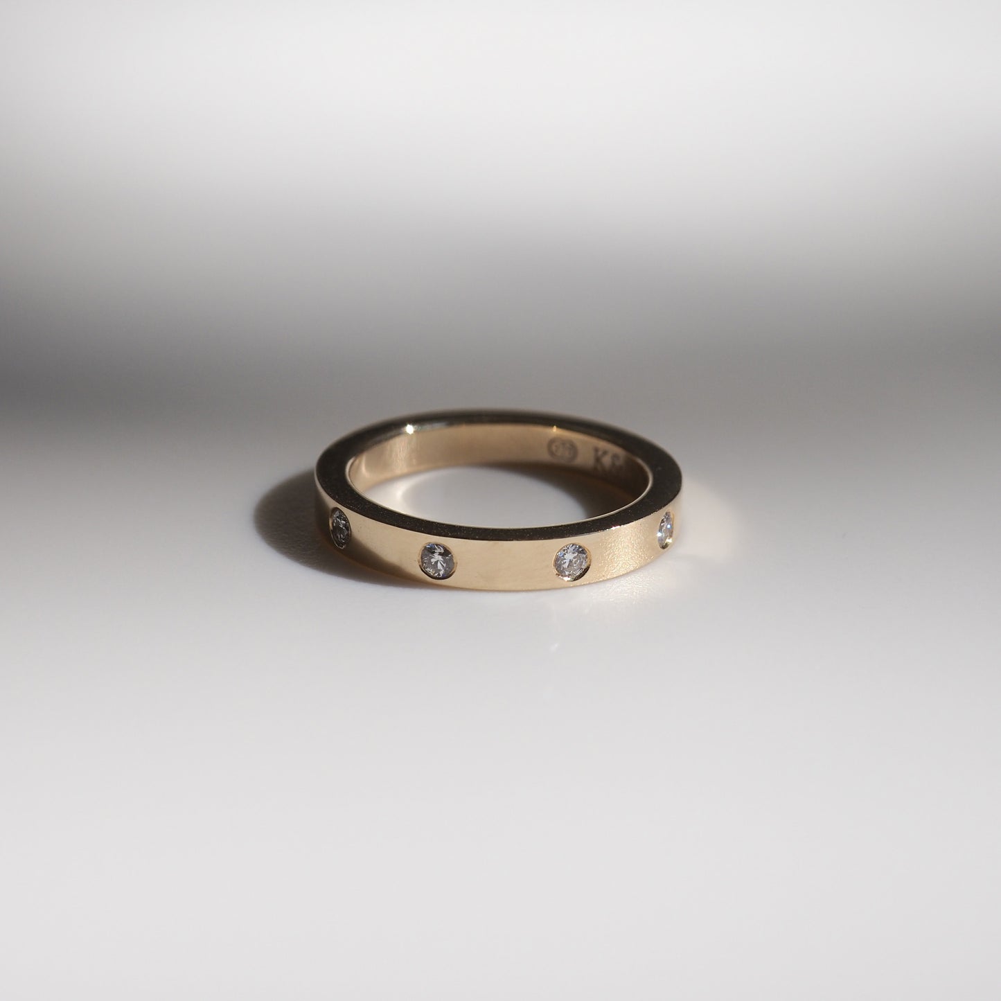 Elegant gold ring with small diamonds evenly spaced around the band, set against a clean, reflective grey surface. Ideal for luxury jewellery lovers.