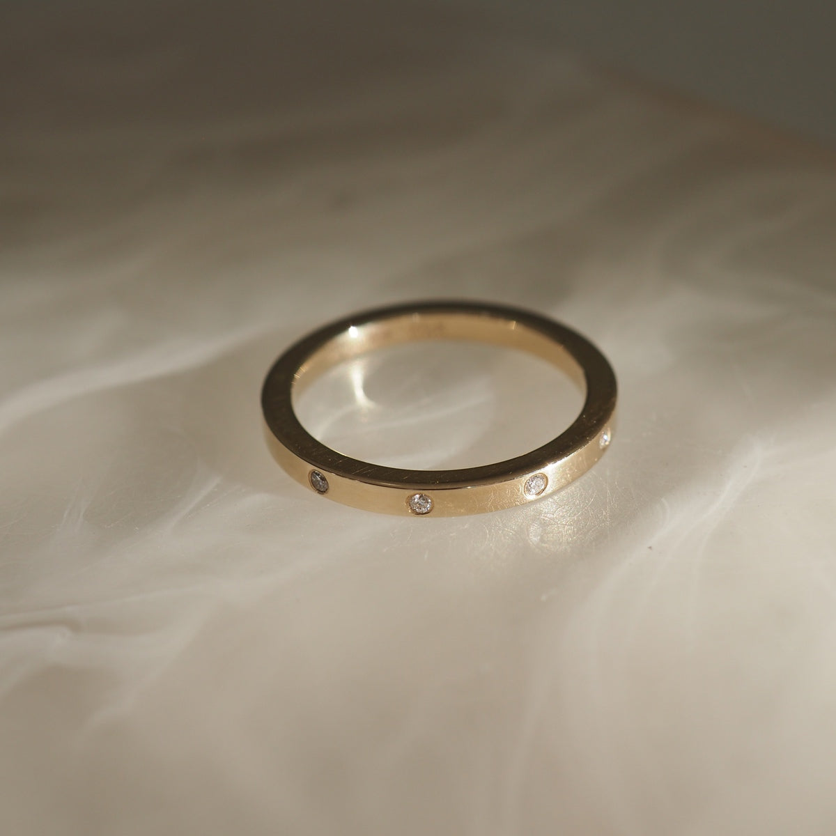 Gold ring with small diamonds set evenly around the band, displayed on a soft, marbled surface. Luxurious jewellery design.