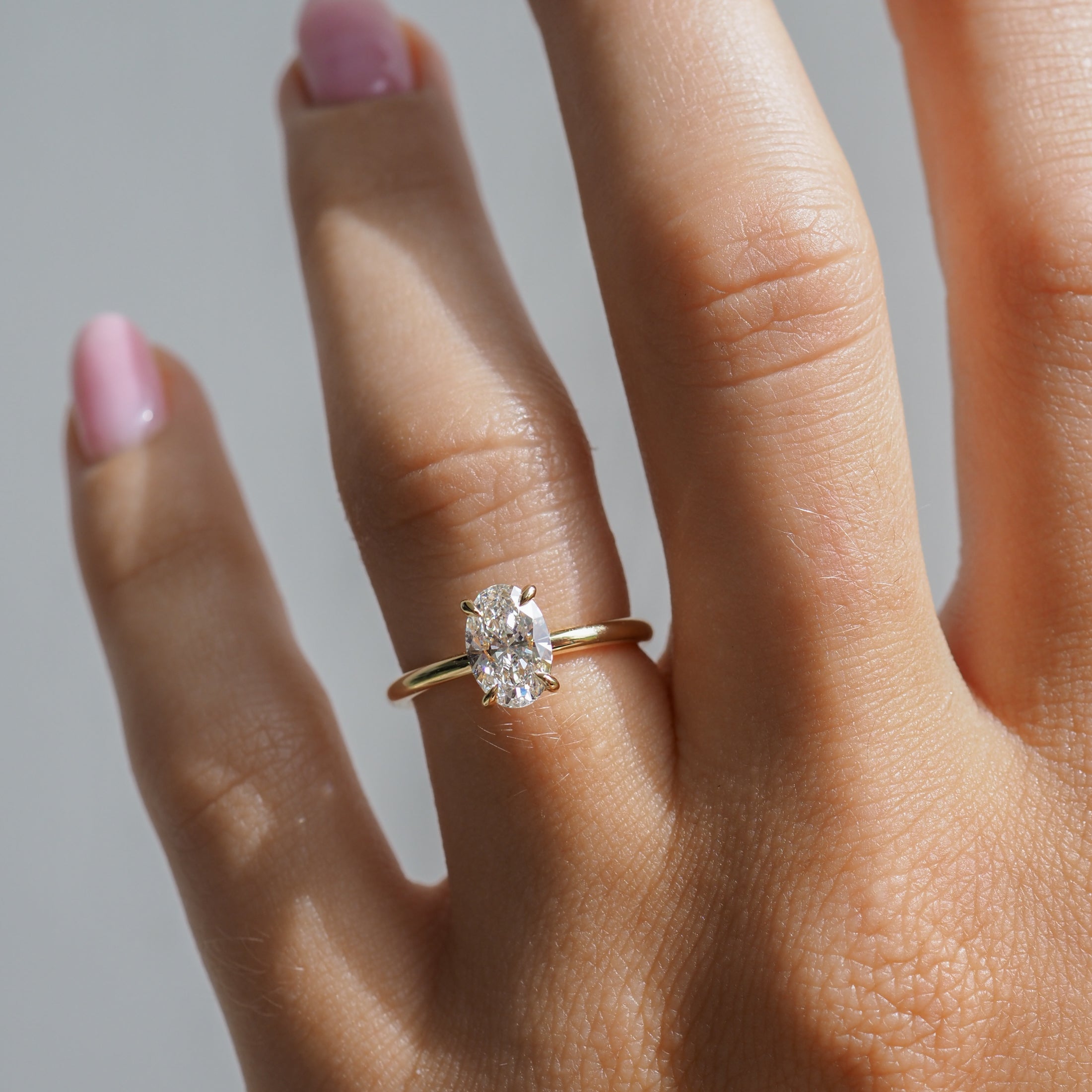 Hand with a gold ring featuring an oval diamond, sunlight highlighting the stone's brilliance. Finger with pastel pink nail polish.