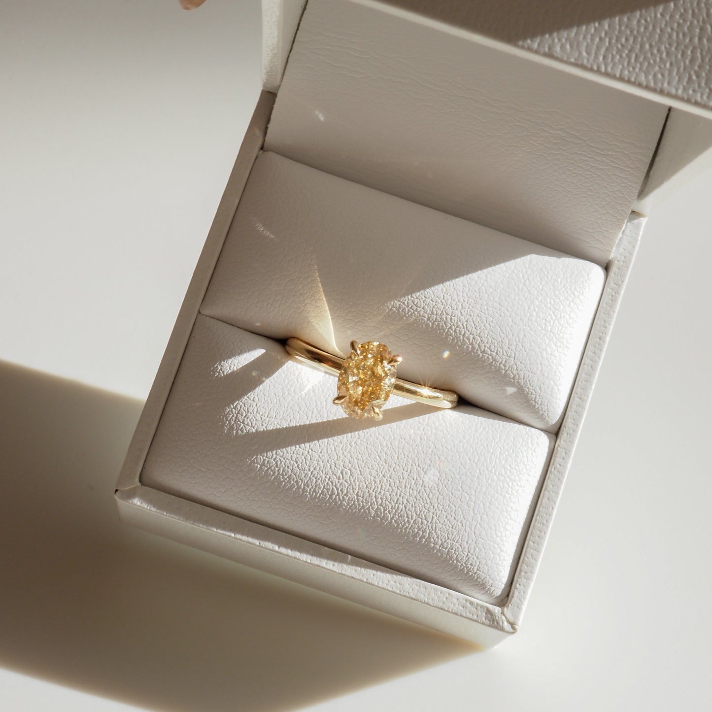 Gold ring with a large, shimmering diamond in a white jewellery box. Sunlight casts delicate shadows, enhancing its luxurious appeal.