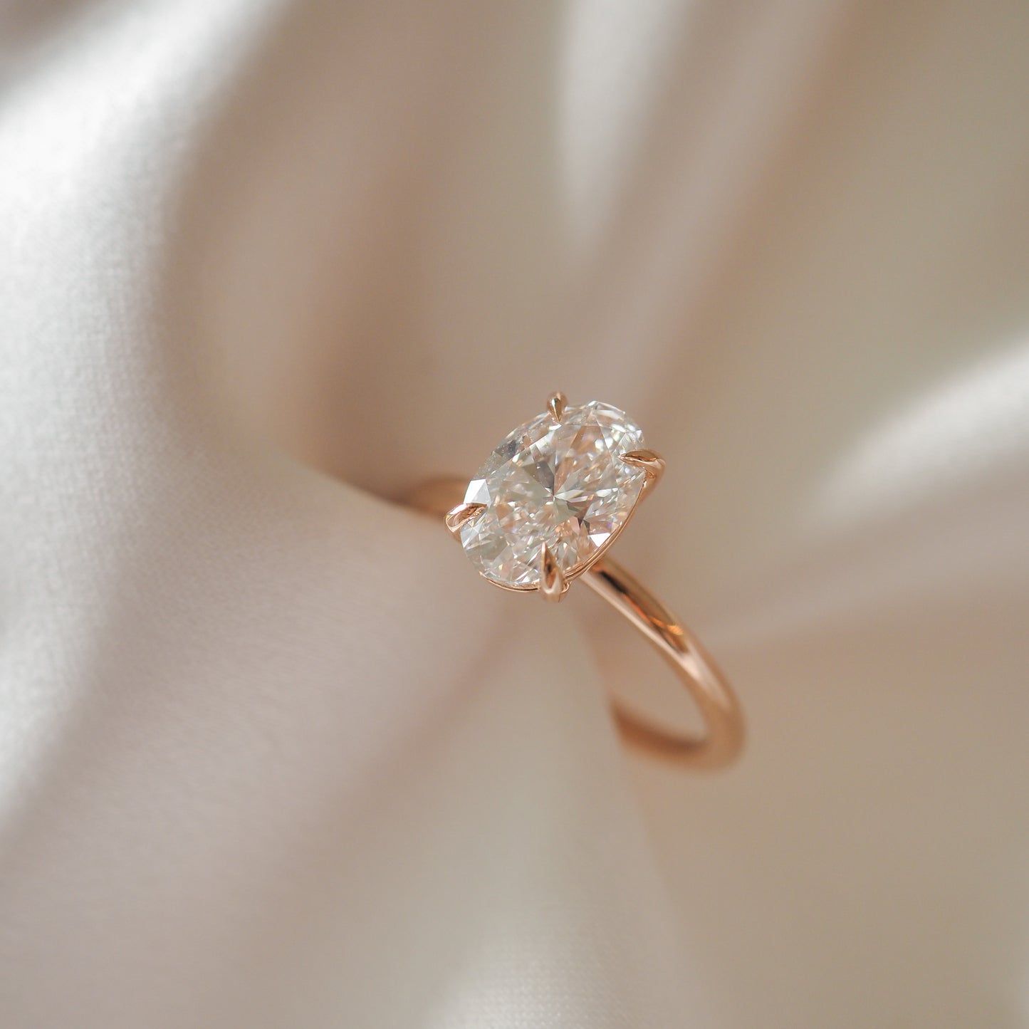 Oval diamond engagement ring with rose gold band on draped white silk; elegant luxury jewellery.
