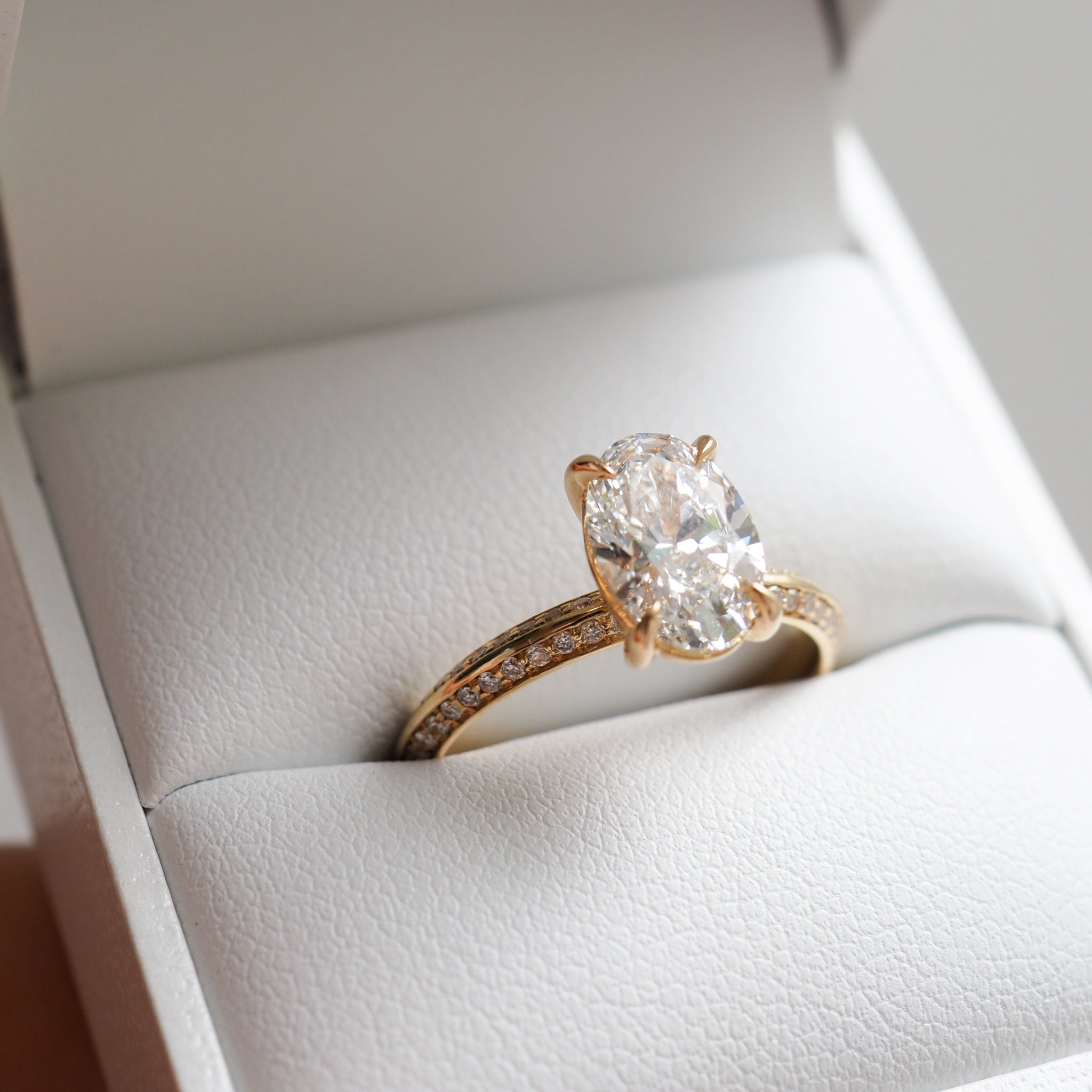 Oval diamond ring in a gold setting, with a pavé band, displayed in an open white jewellery box.