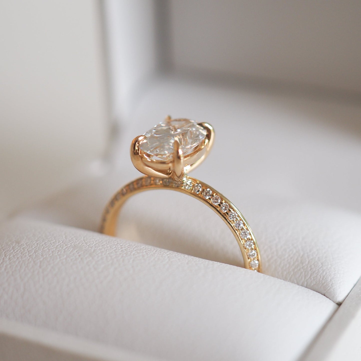 Elegant gold ring with a large oval diamond in a prong setting, featuring pavé diamonds on the band, displayed on a white jewellery box.