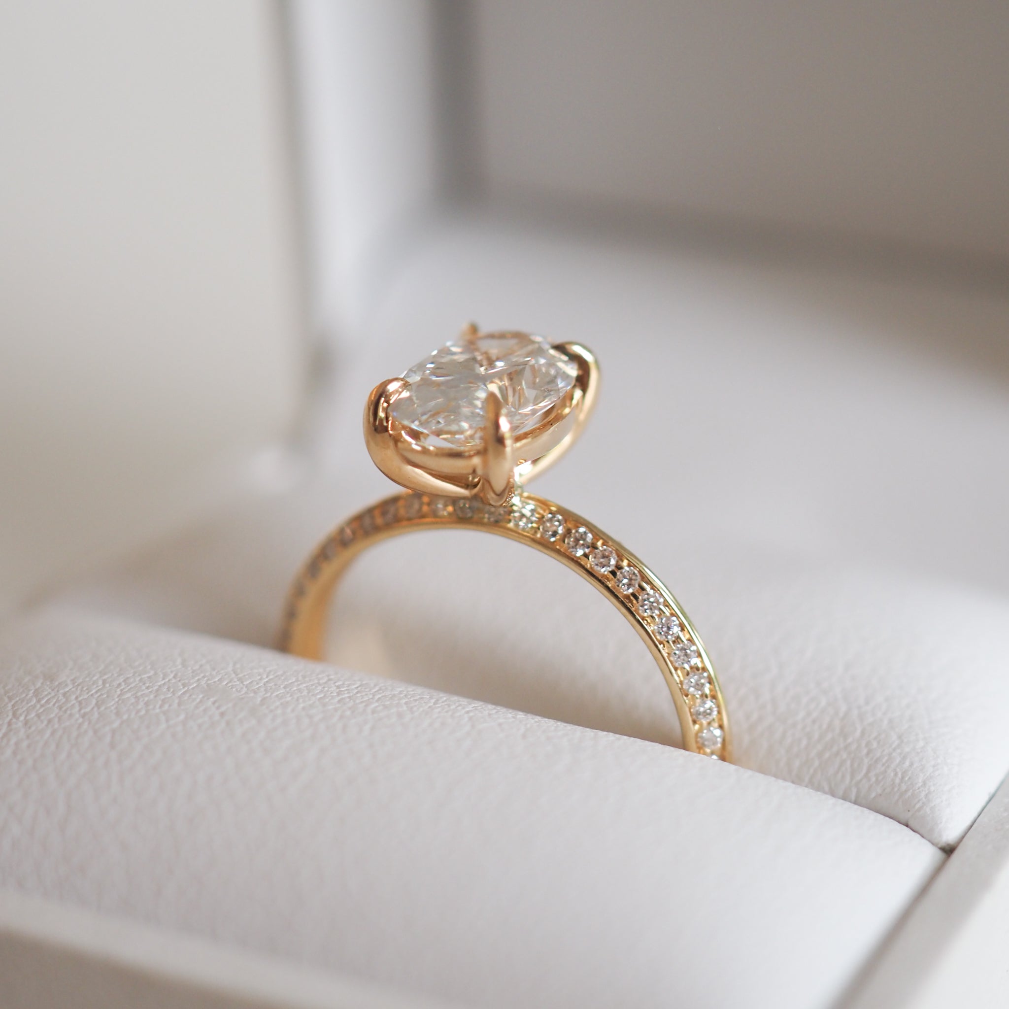 Elegant gold ring with a large oval diamond in a prong setting, featuring pavé diamonds on the band, displayed on a white jewellery box.