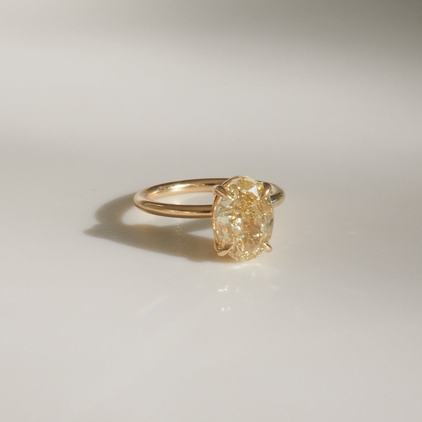 Gold ring with a large oval yellow diamond set in a prong setting, casting a subtle shadow on a plain surface. Luxurious jewellery piece.