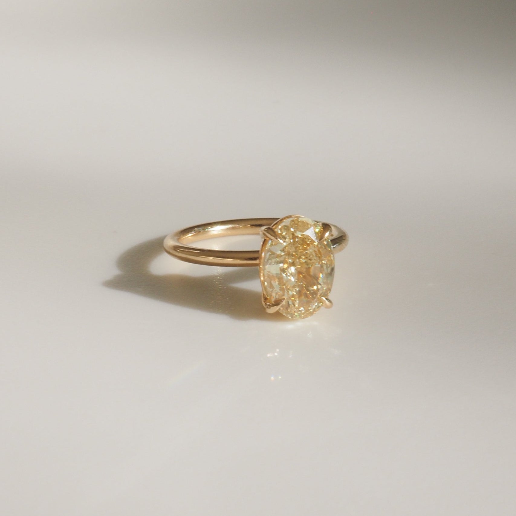 Gold ring with a large oval yellow diamond set in a prong setting, casting a subtle shadow on a plain surface. Luxurious jewellery piece.