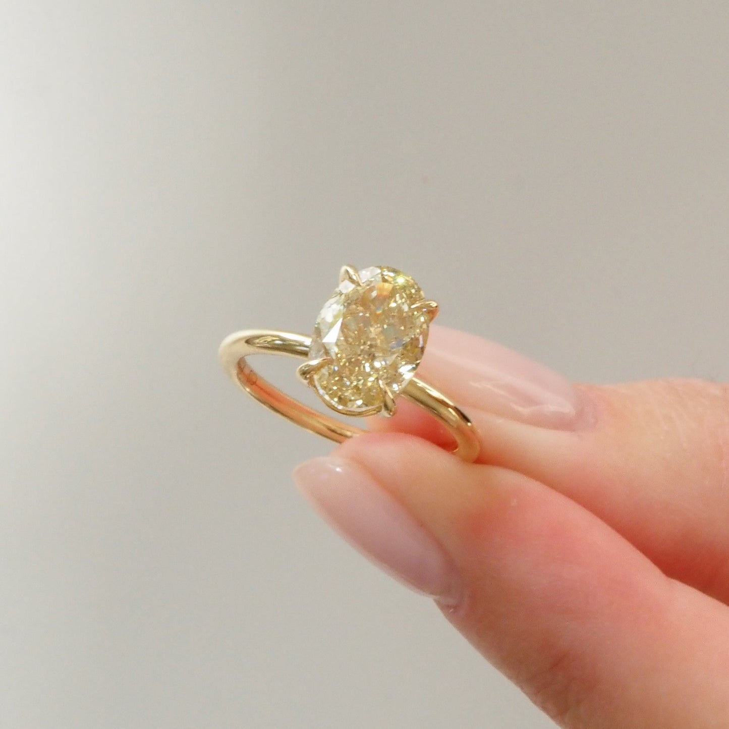 Gold engagement ring with an oval-cut diamond held between two fingers. Elegant luxury jewellery on a neutral background.