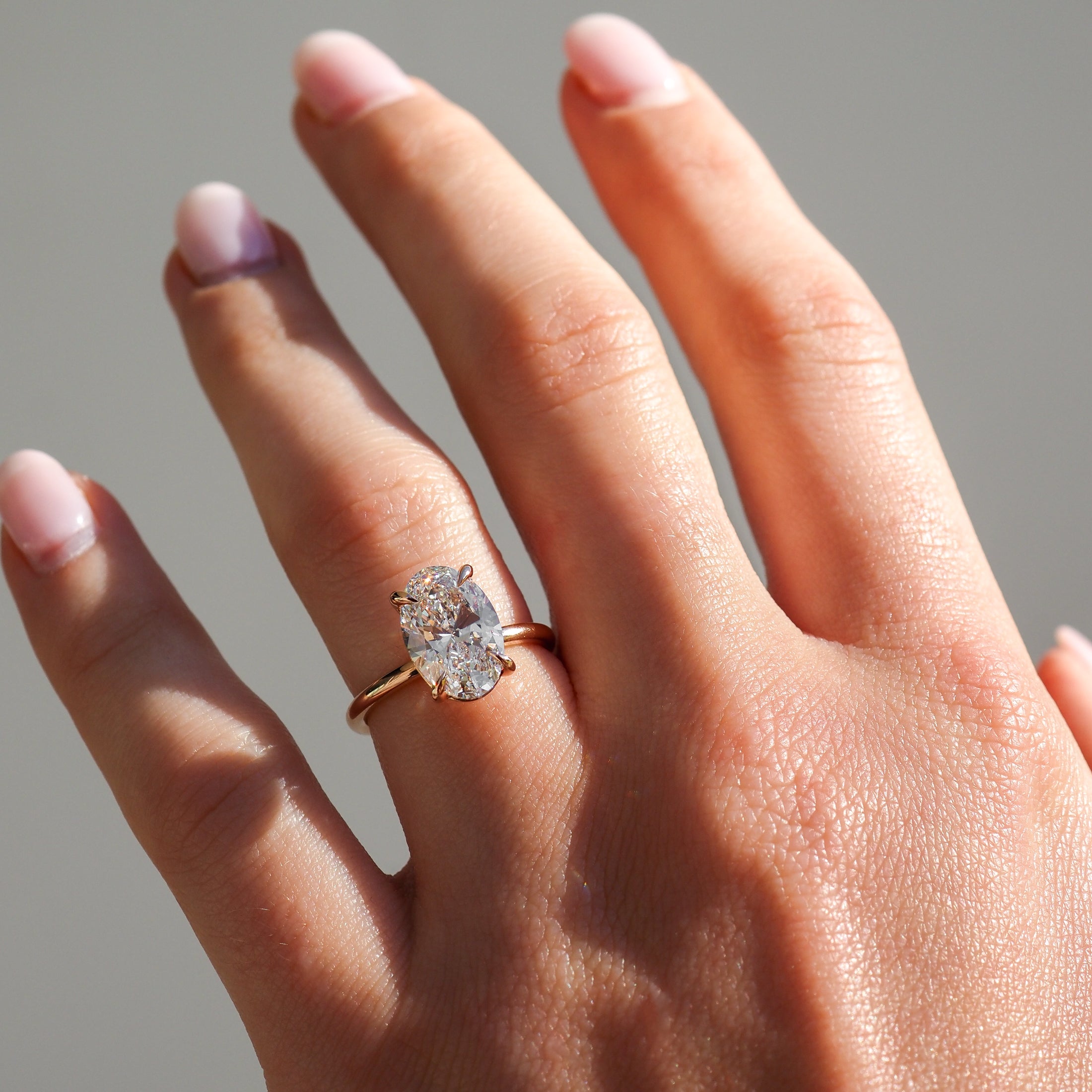 Hand wearing an elegant gold ring with a large oval diamond. The fingers have neatly manicured, pale pink nails, against a neutral background.