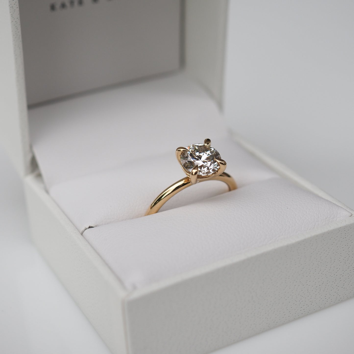 A gold engagement ring with a large, round diamond in a white jewellery box. Ideal luxury gift or proposal accessory.