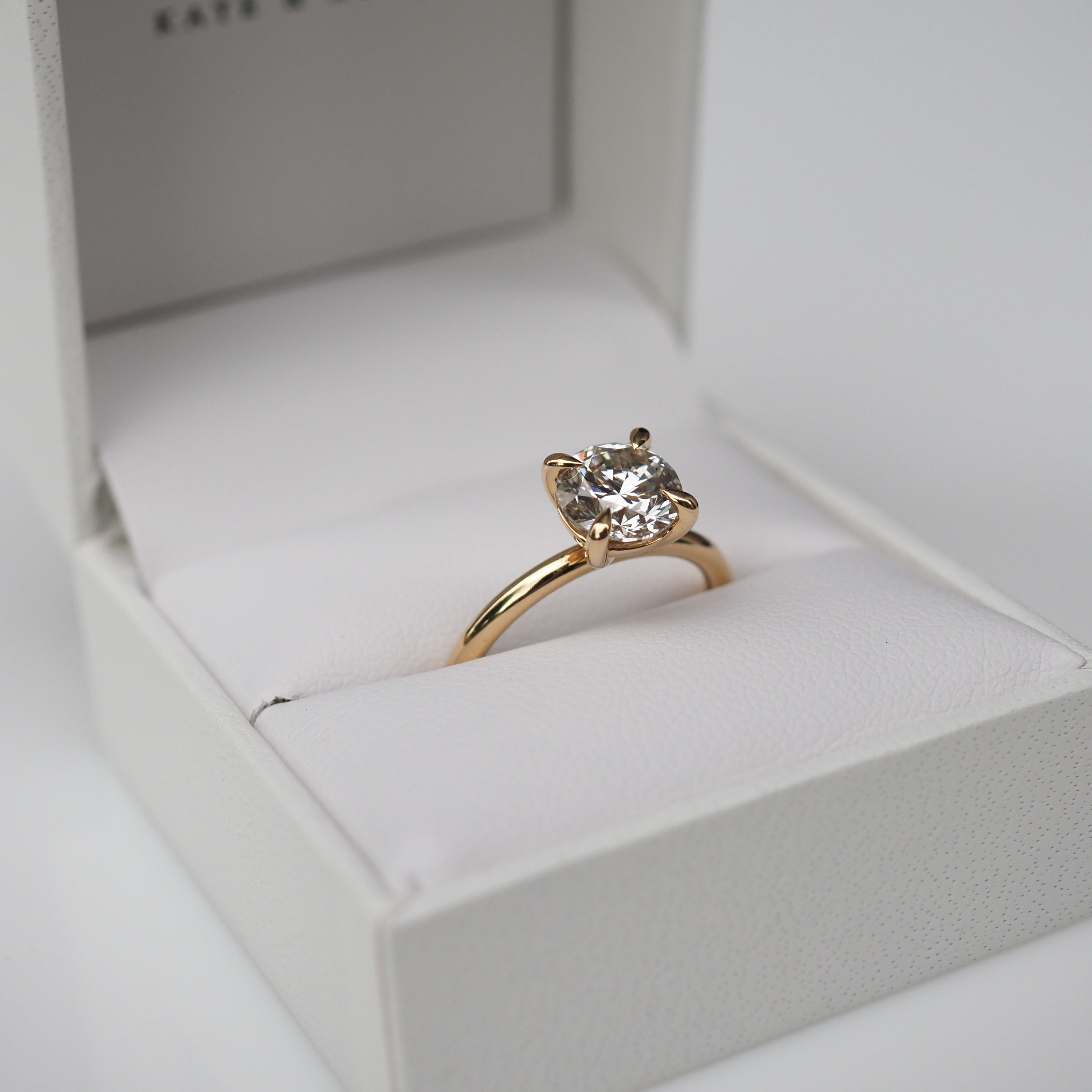 A gold engagement ring with a large, round diamond in a white jewellery box. Ideal luxury gift or proposal accessory.