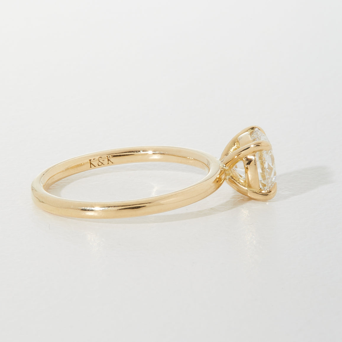 Gold engagement ring with solitaire diamond in a prong setting. Engraved with "K&K," placed on a light background. Luxurious jewellery.