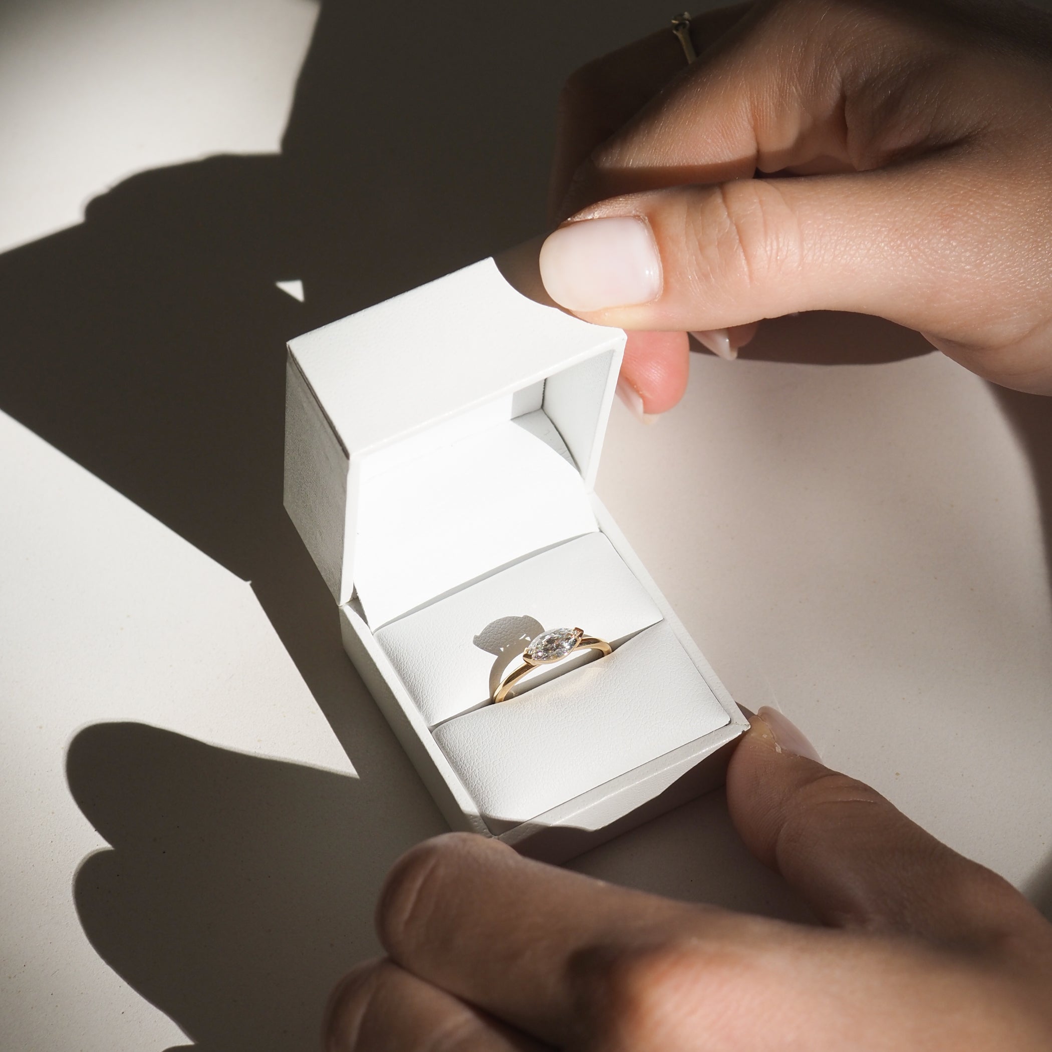 Hands opening a white jewellery box revealing a gold engagement ring with a large diamond, illuminated by soft sunlight.