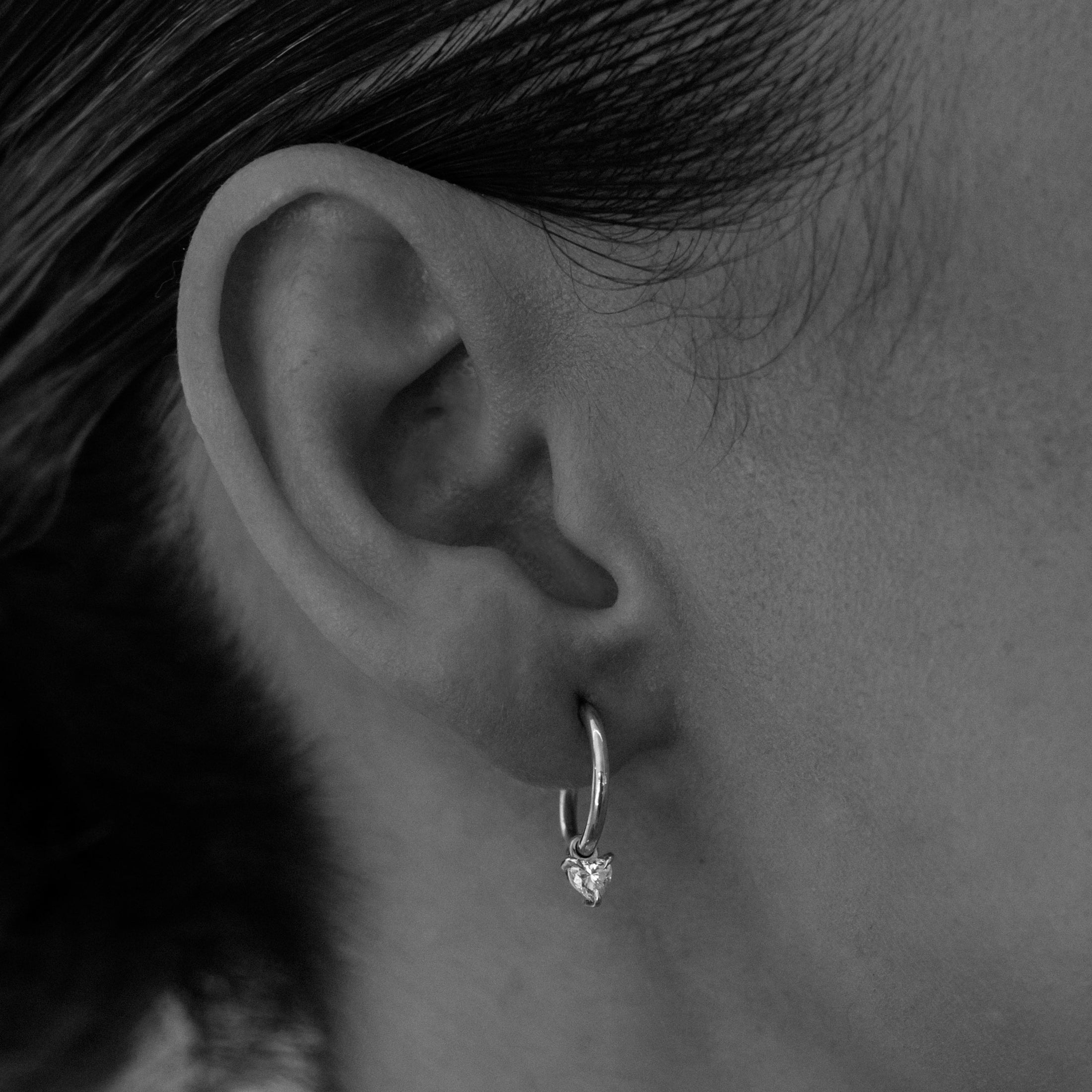 Close-up of an ear wearing a small, elegant hoop earring with a heart-shaped charm. Sleek luxury jewellery in a black and white photo.