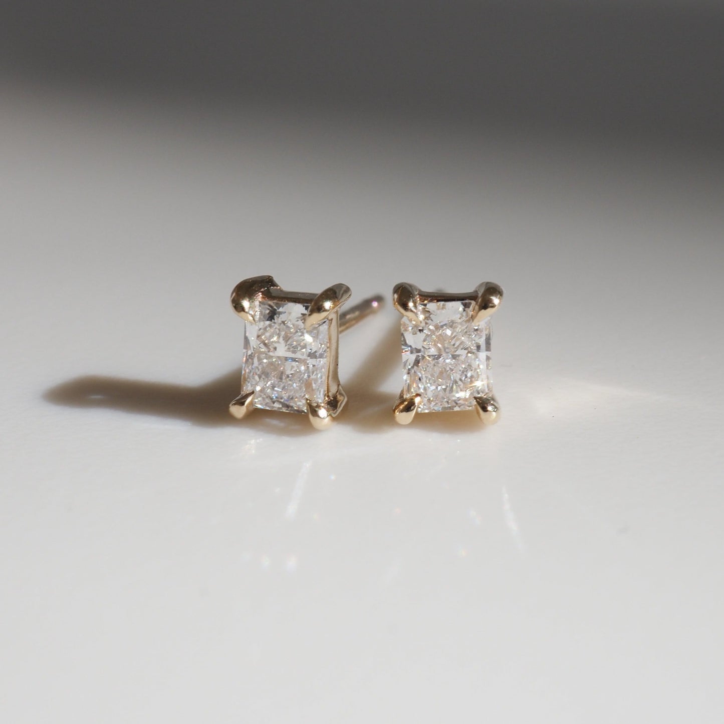 Elegant gold stud earrings with radiant, rectangular-cut diamonds, casting subtle shadows on a smooth surface. Luxury jewellery.