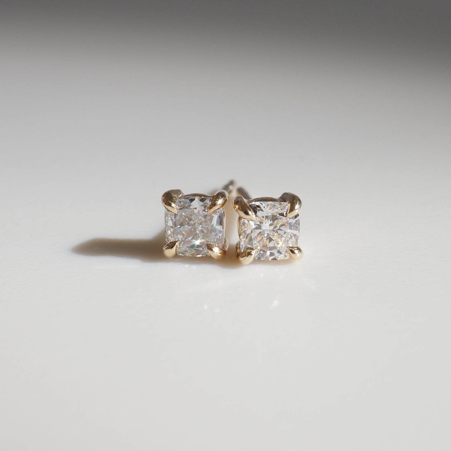 Two princess-cut diamond stud earrings set in gold, elegantly displayed on a smooth, white surface, casting subtle shadows.