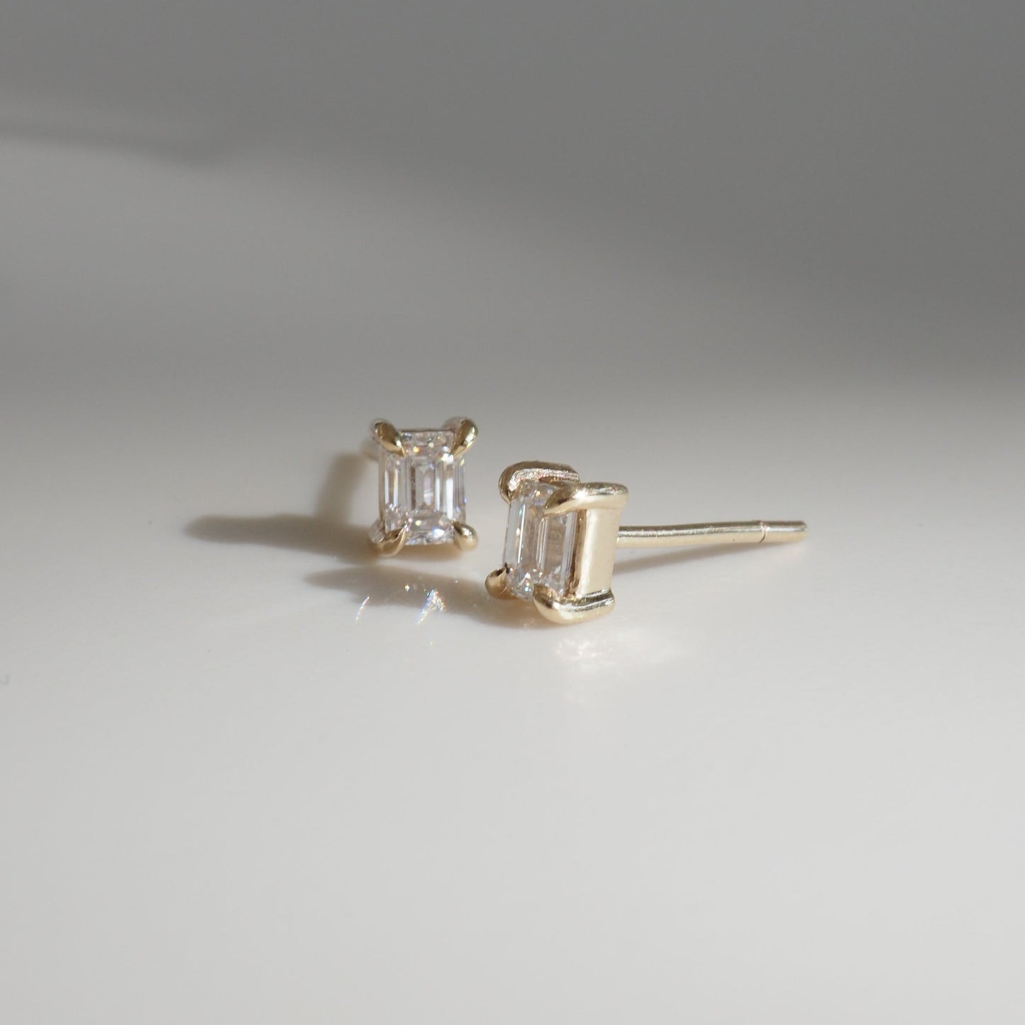Emerald-cut diamond stud earrings set in gold, displayed on a smooth surface under soft lighting. Luxurious fine jewellery.