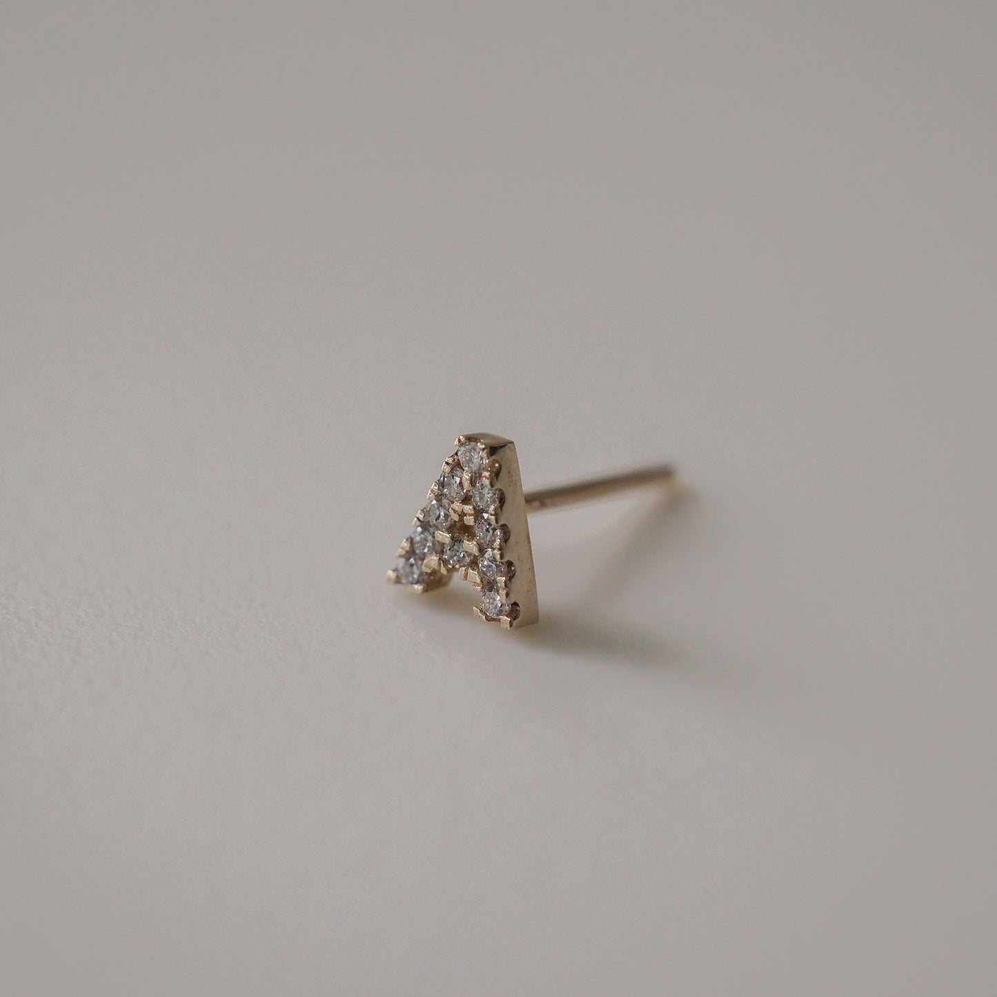 Gold stud earring shaped like the letter 'A', encrusted with sparkling diamonds, set against a soft grey background.