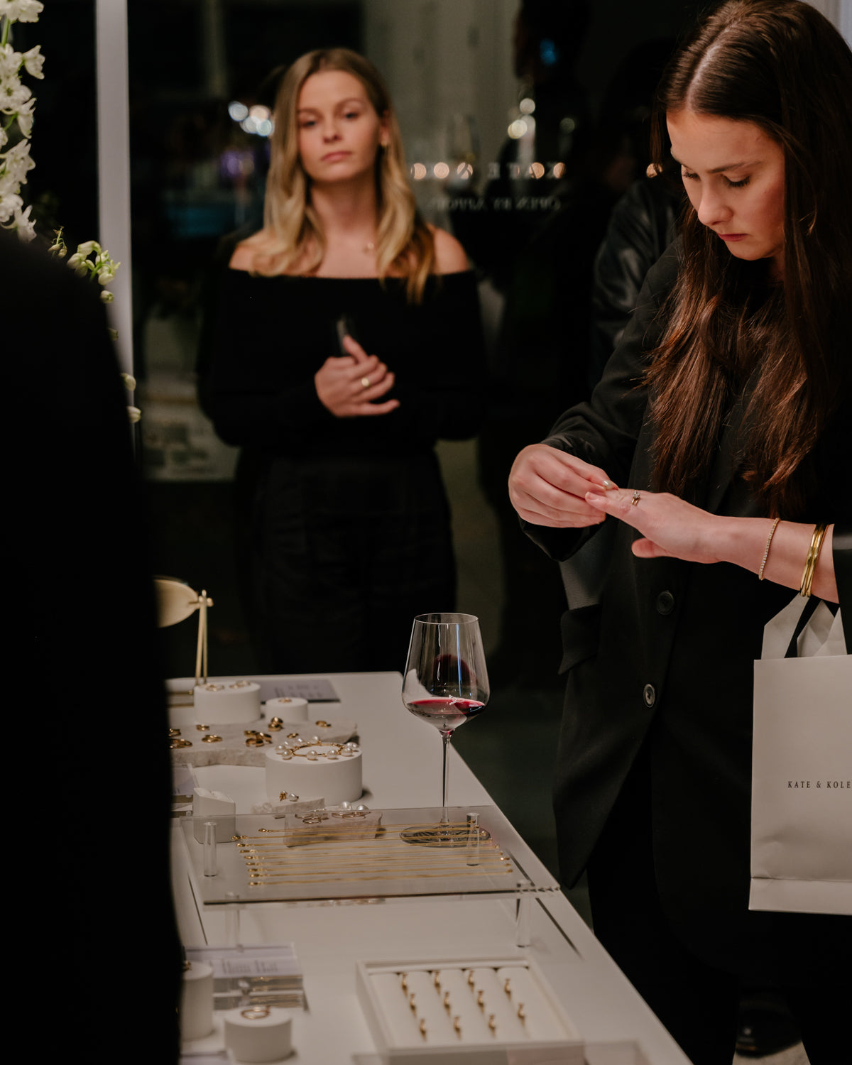 A woman examines high-end jewellery at an elegant display, holding a shopping bag. A glass of red wine sits nearby; another woman observes.
