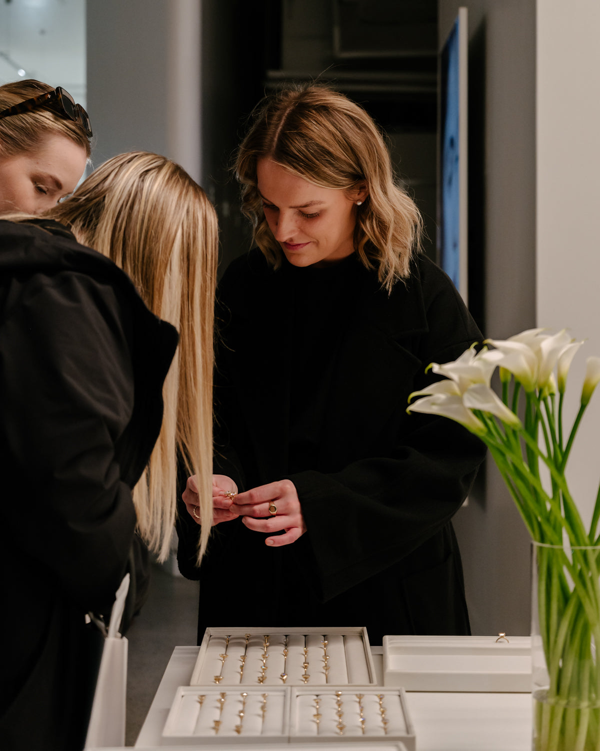 Two women admire gold rings in a luxury jewellery store. A table displays elegant rings, with white flowers in a vase beside them.