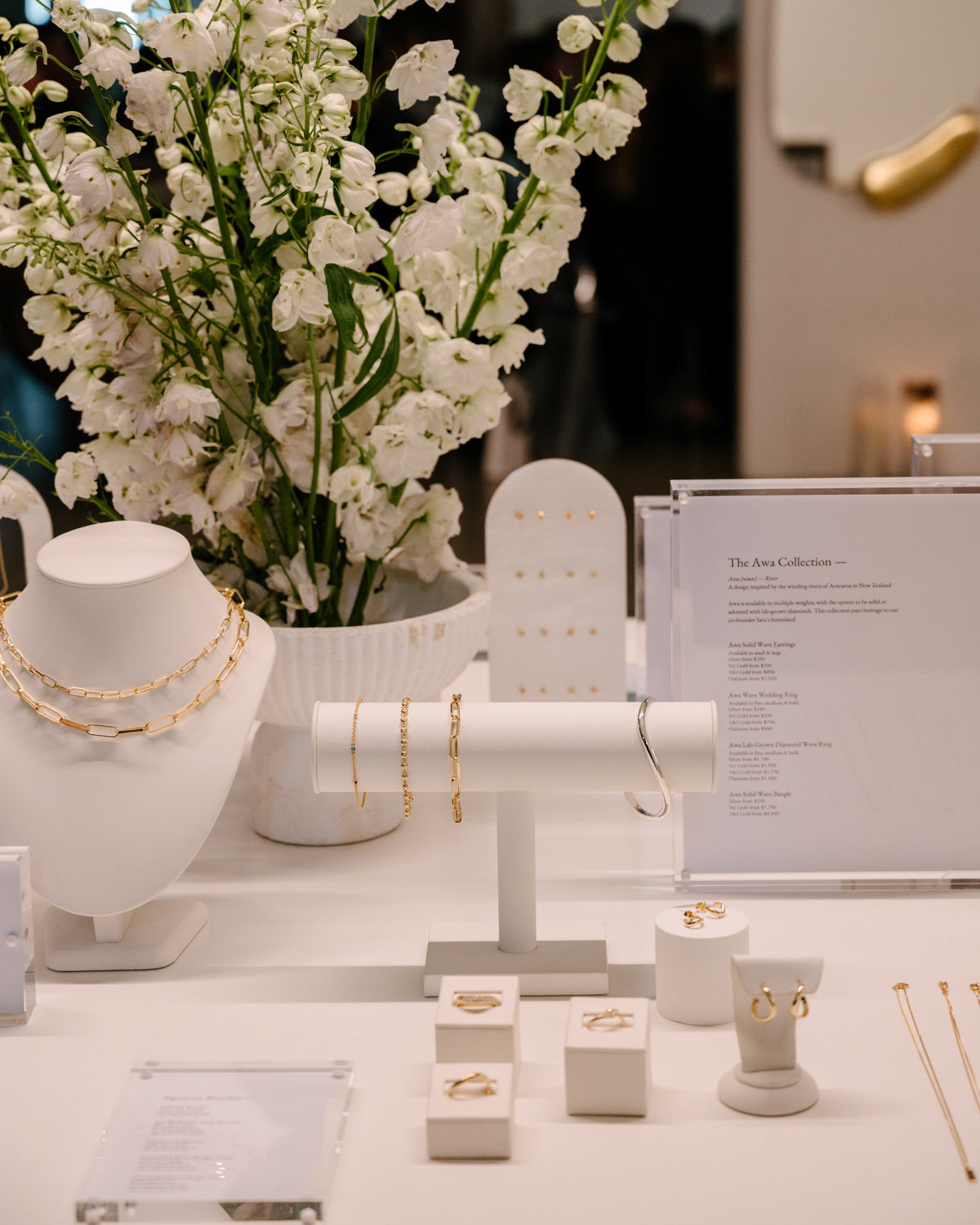 Elegant jewellery display featuring gold necklaces, bracelets, and rings on white stands; lush white floral arrangement in the background.