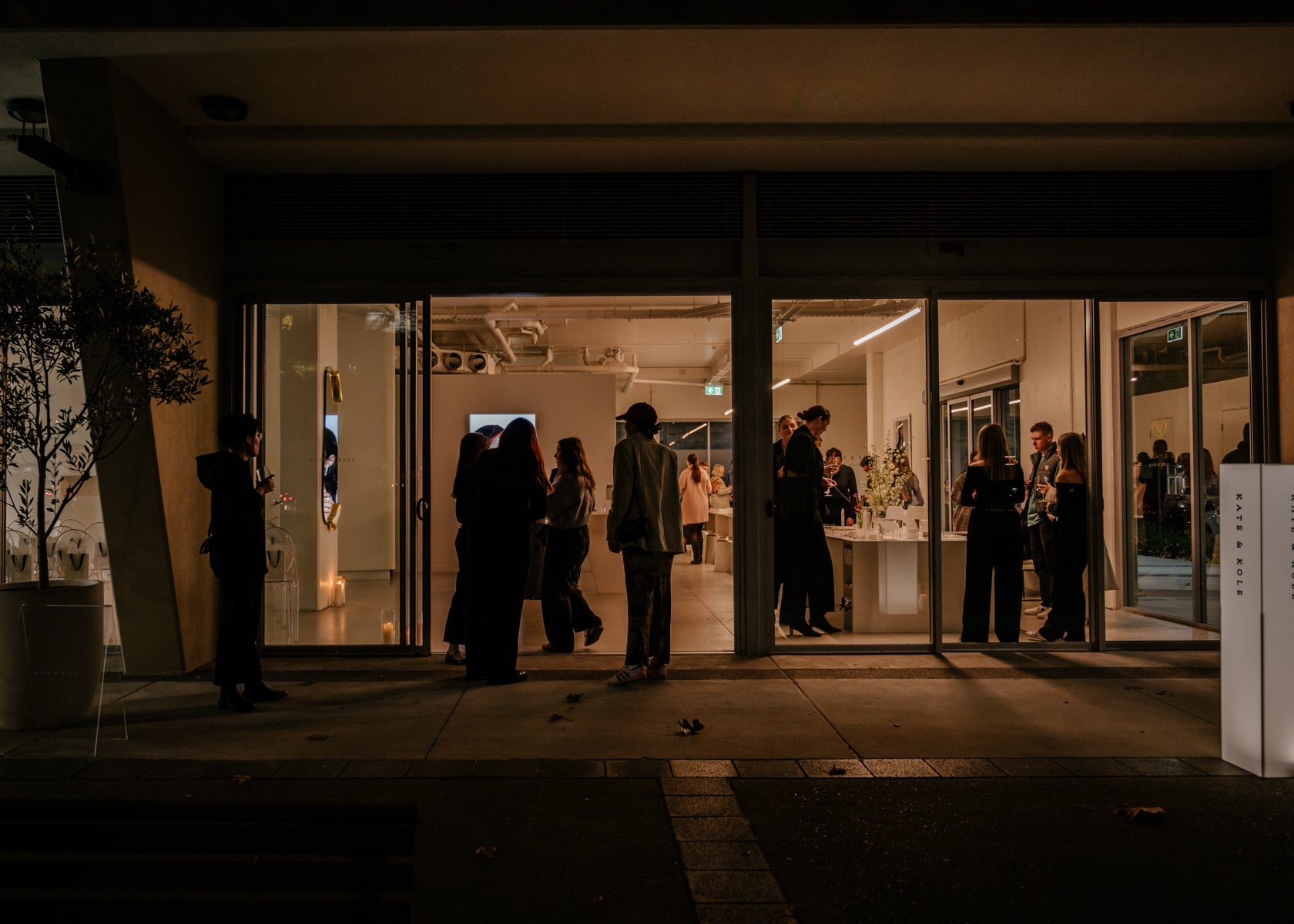 Elegant jewellery store event at night, attendees socialising inside. Warm interior lighting, modern decor, glass windows showcasing luxury pieces.