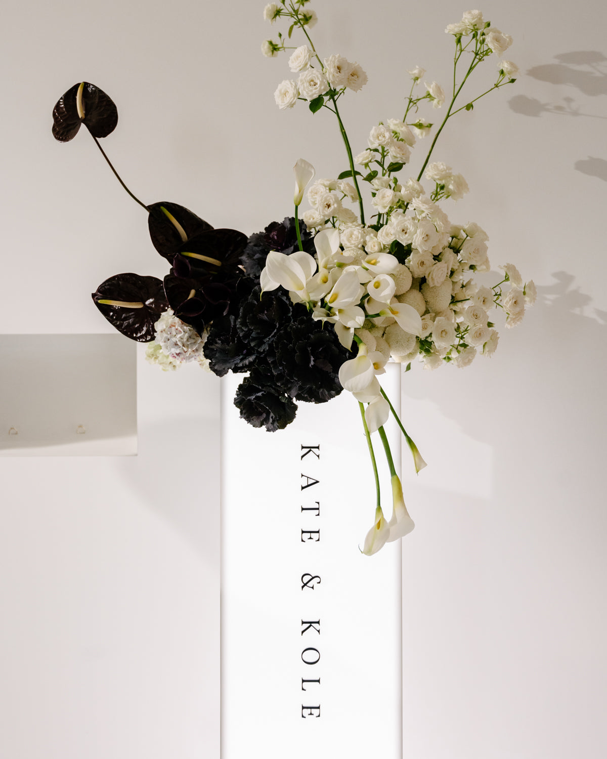 Floral arrangement with white blooms and dark leaves on a minimalist background, featuring "KATE & KOLE". Elegant high-end jewellery display.