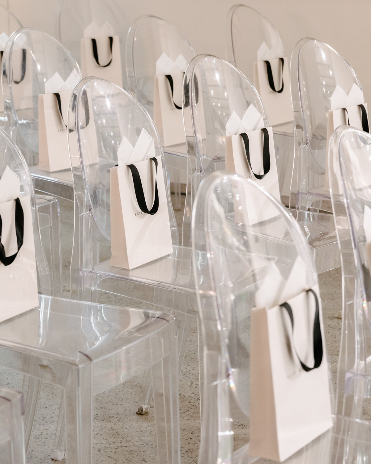 Clear ghost chairs with white gift bags featuring black handles are neatly arranged in rows on a polished concrete floor.