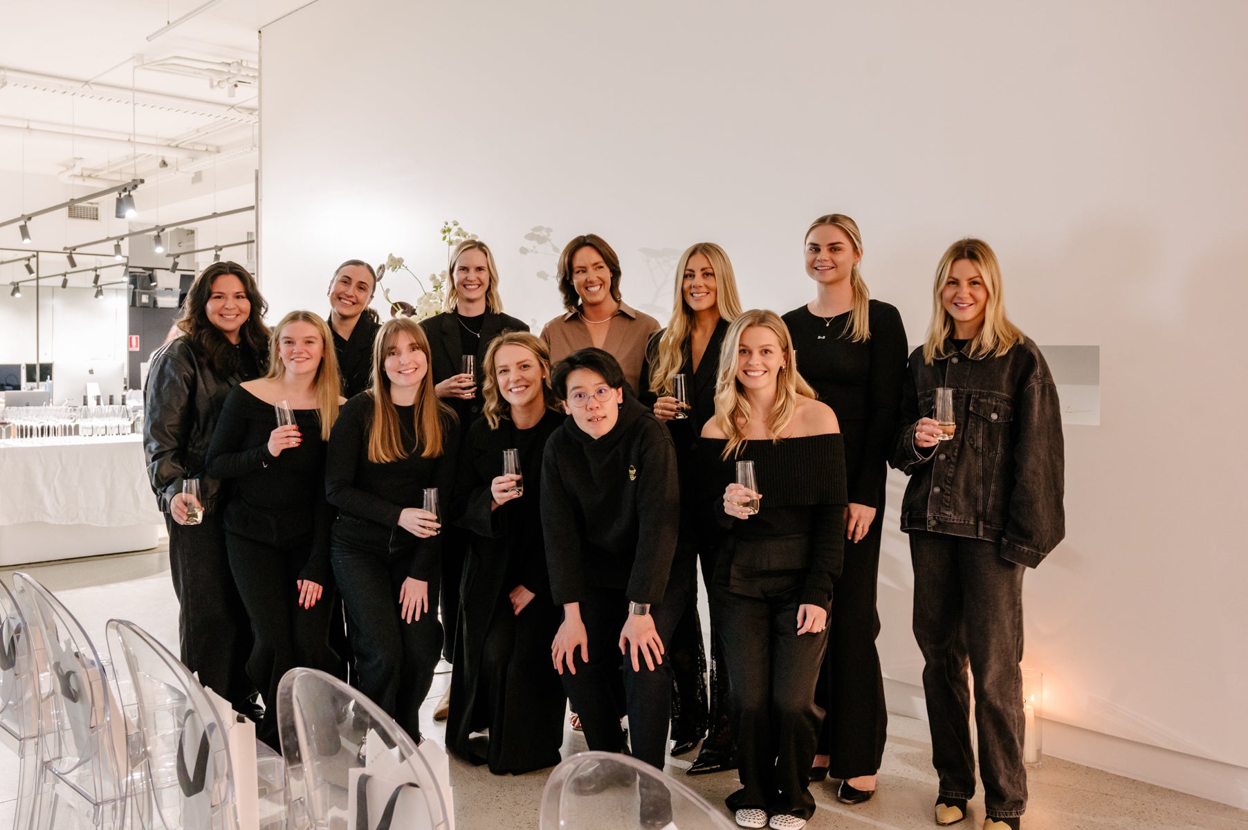 A group of women in black attire stand smiling in an elegantly lit venue, holding champagne glasses, possibly celebrating a luxury jewellery event.