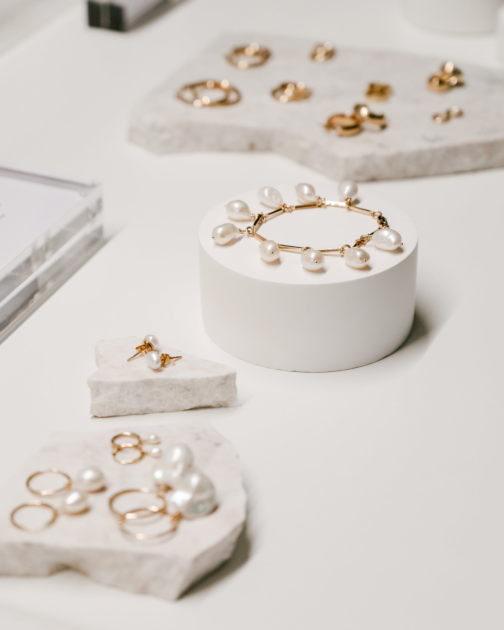 Elegant display of luxury jewellery with gold rings, pearl drop earrings, and a pearl bracelet on white stone stands.