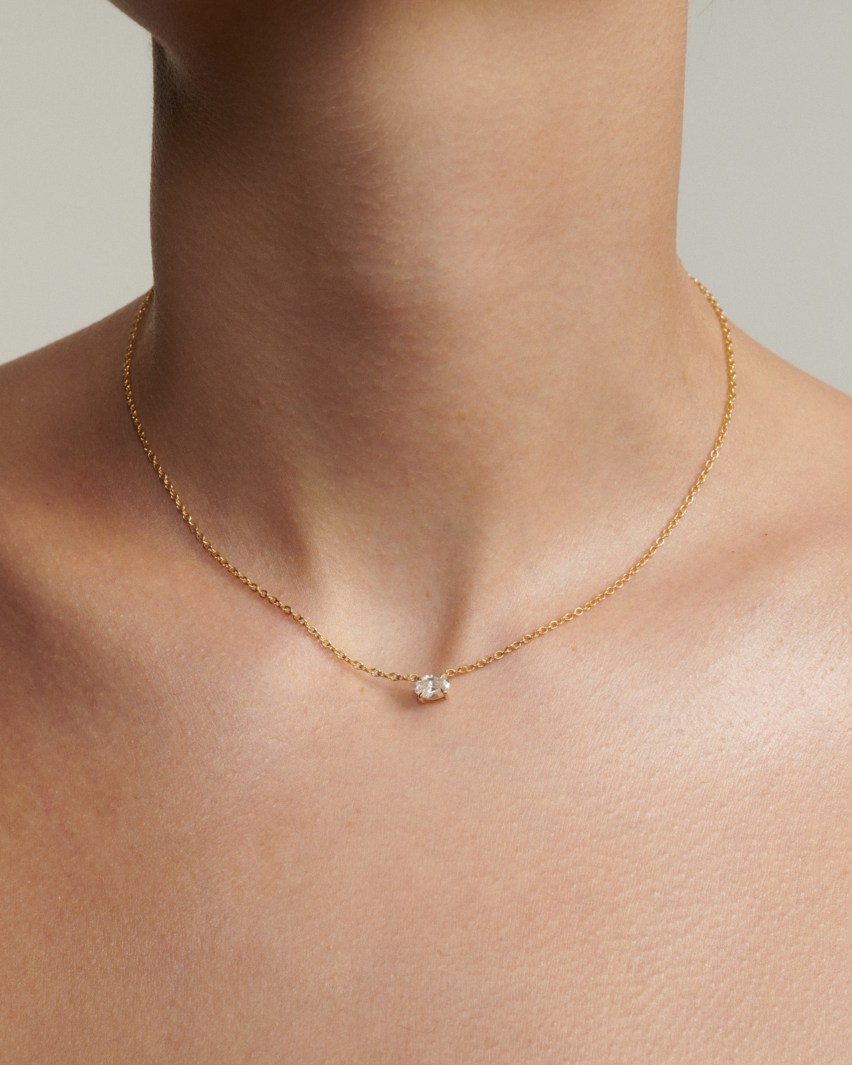 Gold necklace with a diamond pendant on a person's neck against a neutral background [text overlay: Andrea wears the 0.5ct East West Fixed Oval Diamond Necklace]