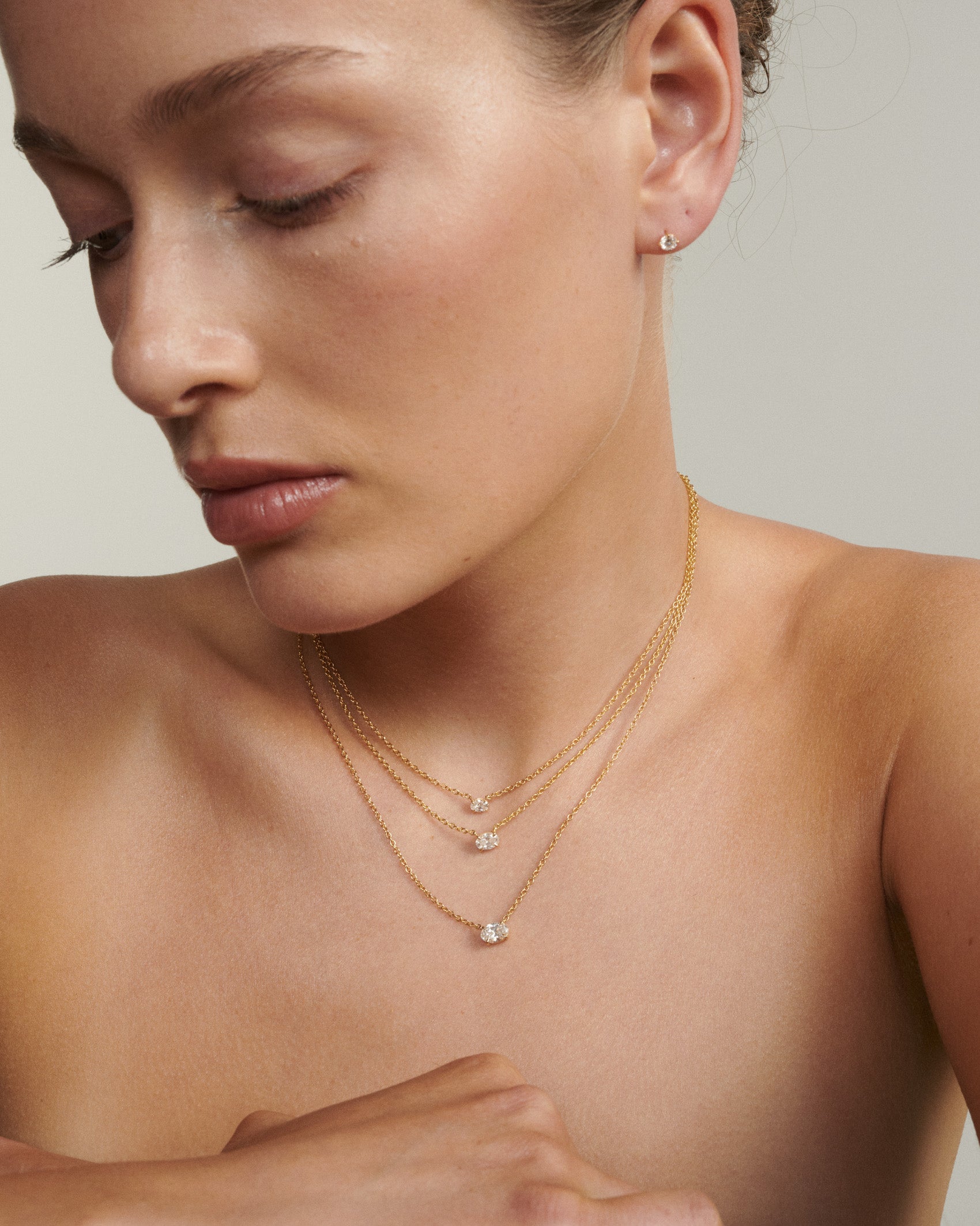 Close-up of a woman wearing gold necklaces and earrings on a neutral background [text overlay: Andrea wears the 0.25, 0.5ct & 0.75ct East West Fixed Oval Diamond Necklace]