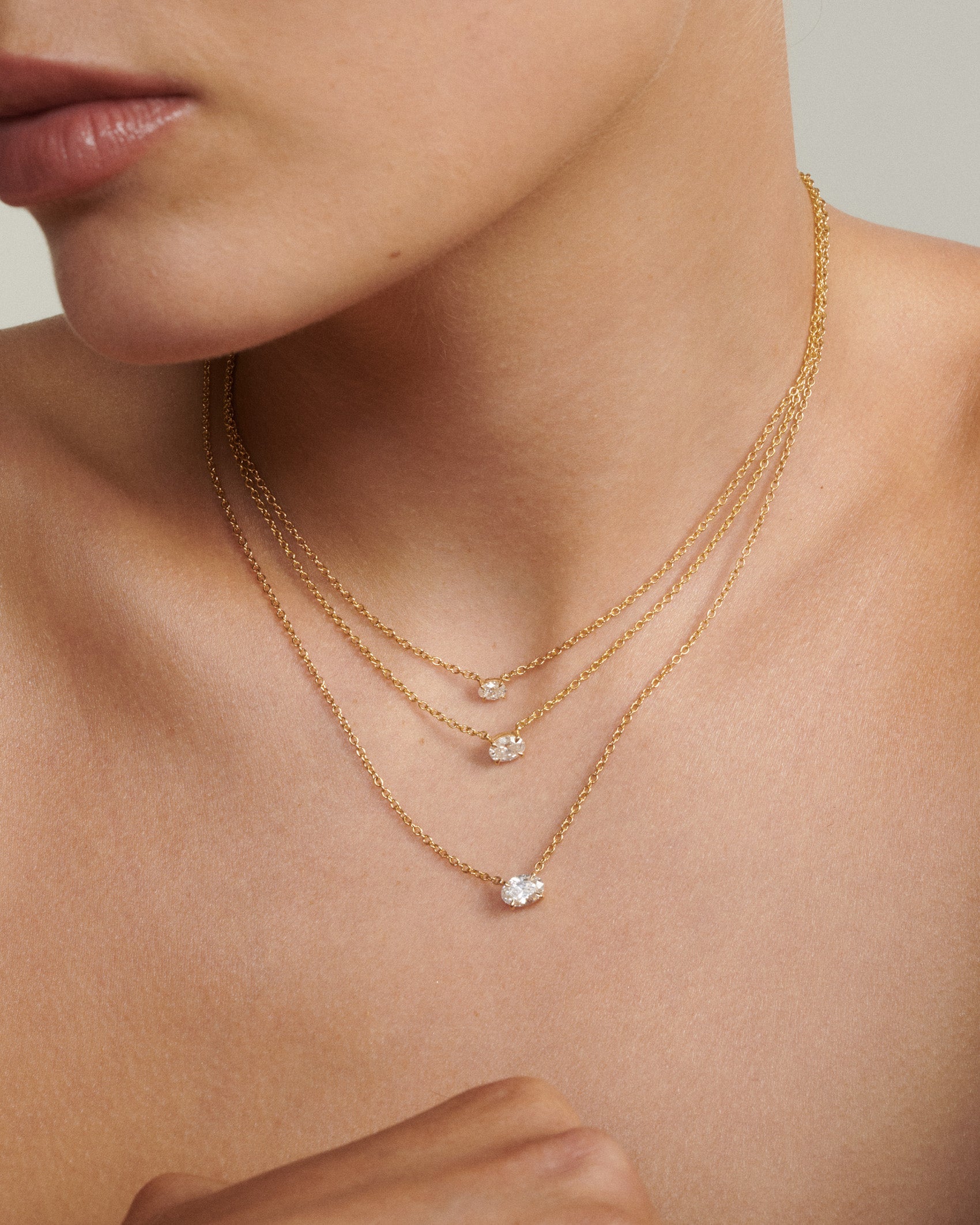 Close-up of a person wearing two gold necklaces with small pendants on a neutral background [text overlay: Andrea wears the 0.25, 0.5ct & 0.75ct East West Oval Diamond Necklace]
