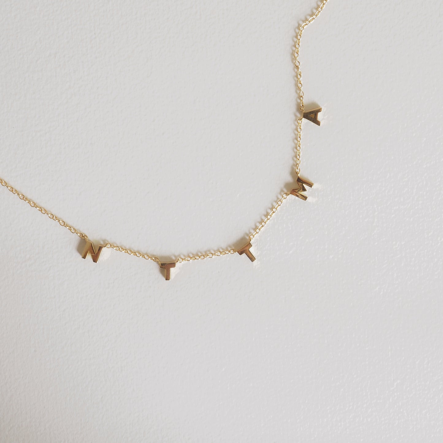 Gold necklace with small, spaced letter charms spelling "WRITE" on a delicate chain, against a light background.