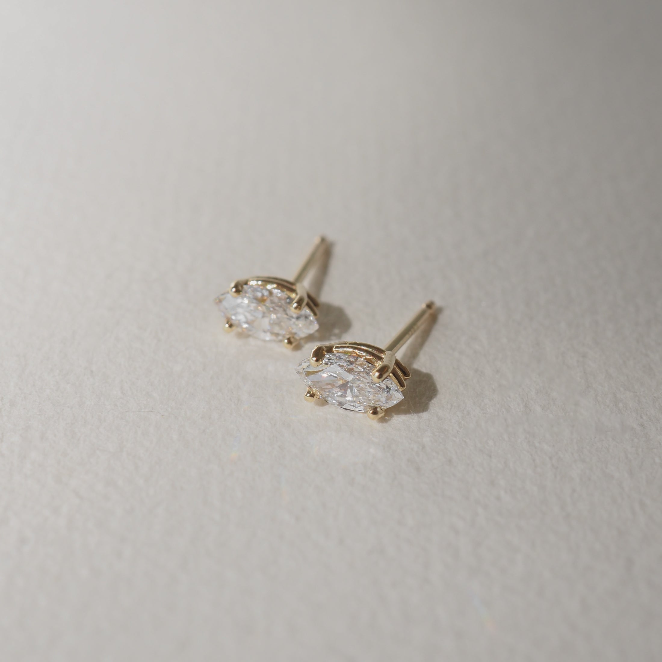 Two marquise-cut diamond stud earrings set in gold, placed on a textured, light background. Ideal for luxury jewellery enthusiasts.